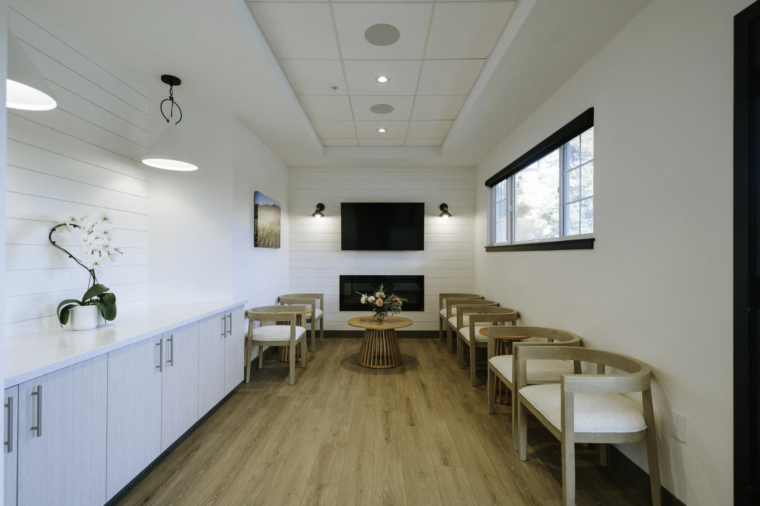A wide view of the waiting room shows the beverage station accented with shiplap siding and farmhouse pendants from Visual Comfort. Also featured is a tray ceiling with acoustic ceiling panels and recessed lighting.