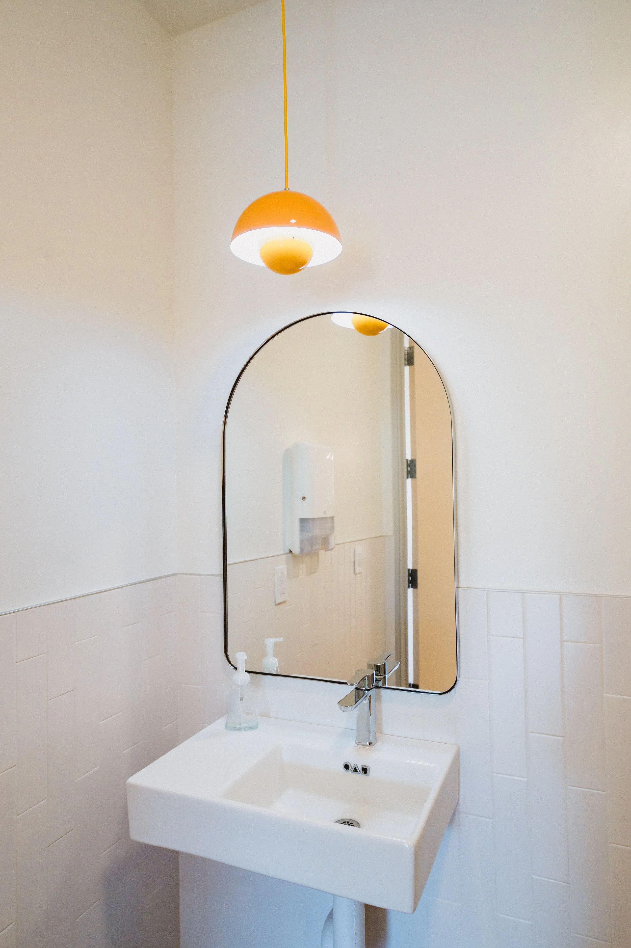 A bathroom with a white sink, a mirror, a soap dispenser, and a hanging light fixture.
