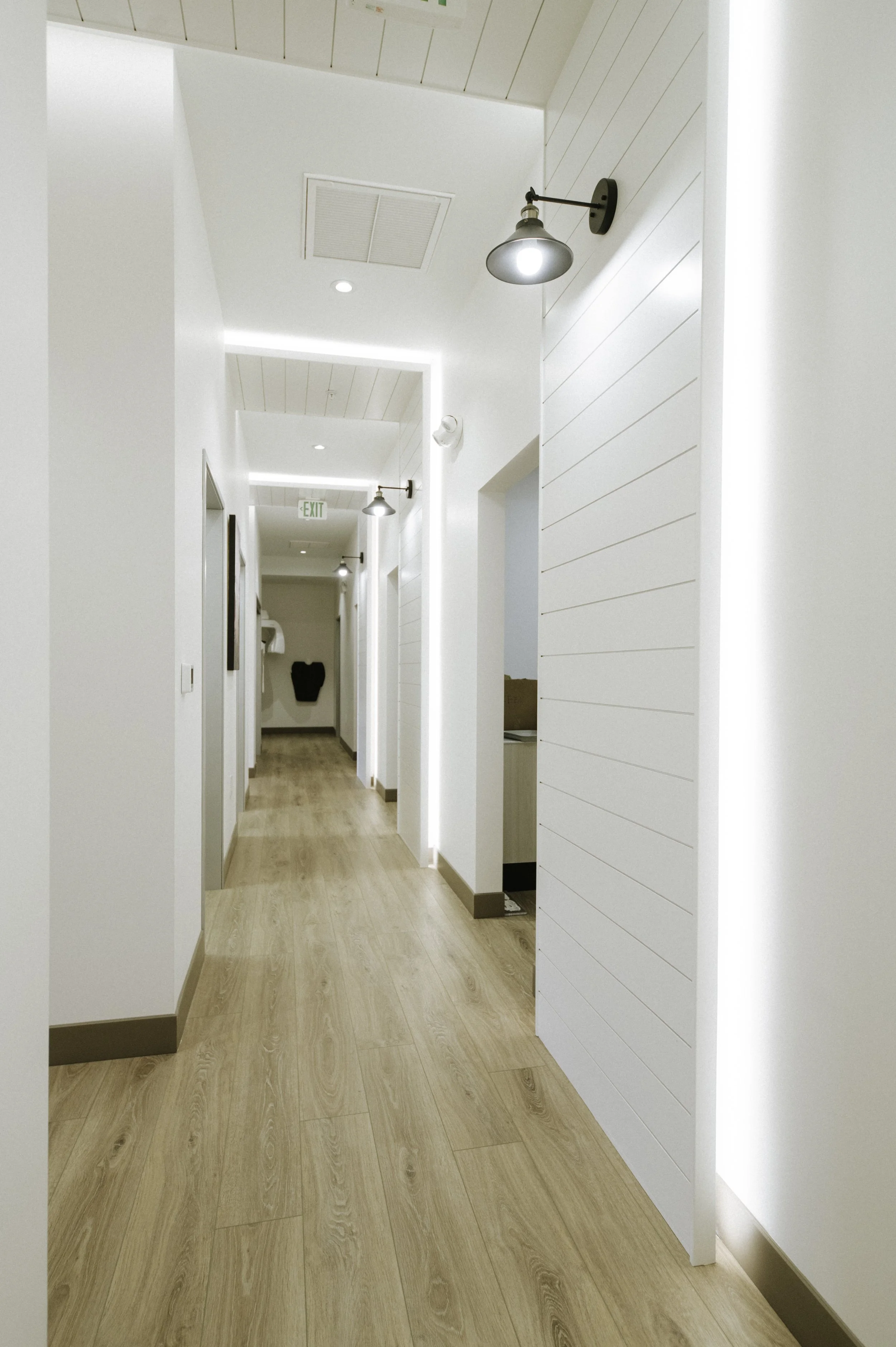 Warm LVT flooring is trimmed with black moulding. Architectural lighting provides wayfinding and sets off the nickle gap shiplap accent walls with farmhouse sconces.