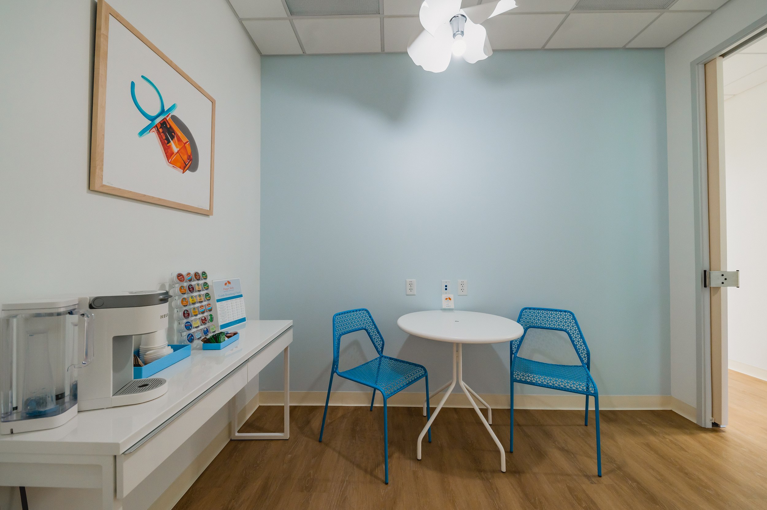 Small indoor space with a white table, two blue chairs, a coffee station with a water dispenser and coffee pods, and a framed abstract artwork on a white wall.