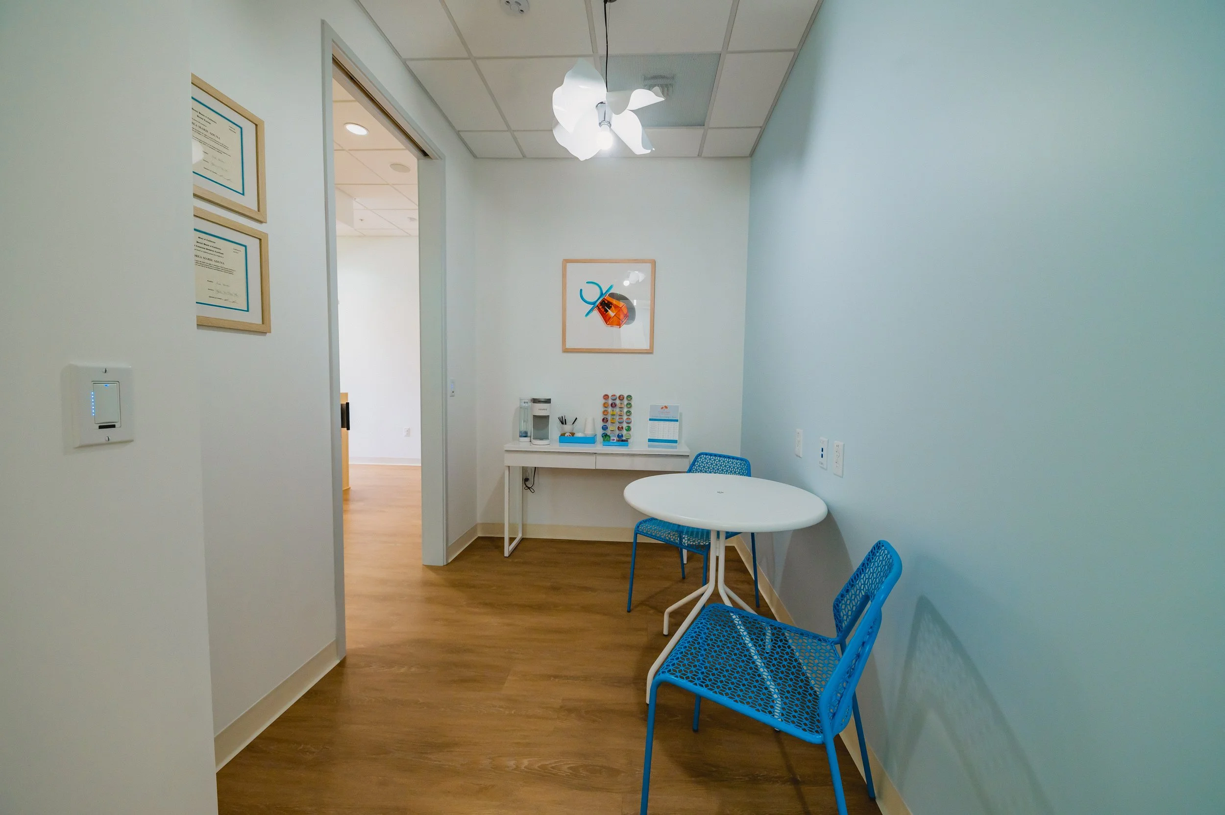 A small area with a white round table and two blue chairs. A white cabinet with a coffee maker and supplies is against the wall, above the table. A framed picture of a ladybug is on the wall. The walls are painted light blue, and the floor is wooden.