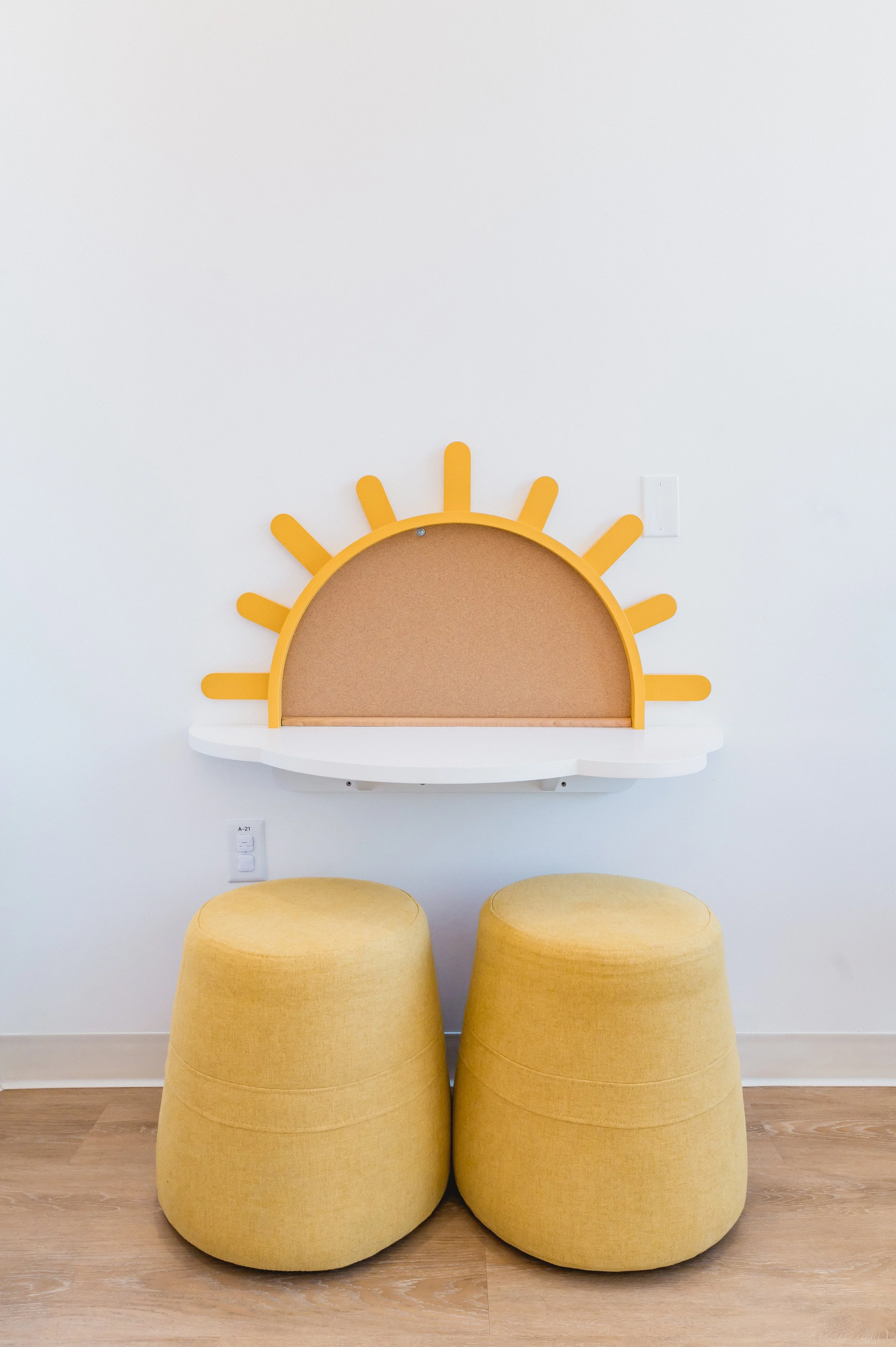 Decorative sun-shaped bulletin board with yellow rays above a white shelf, two yellow rounded ottomans in front, against a white wall and wooden floor.