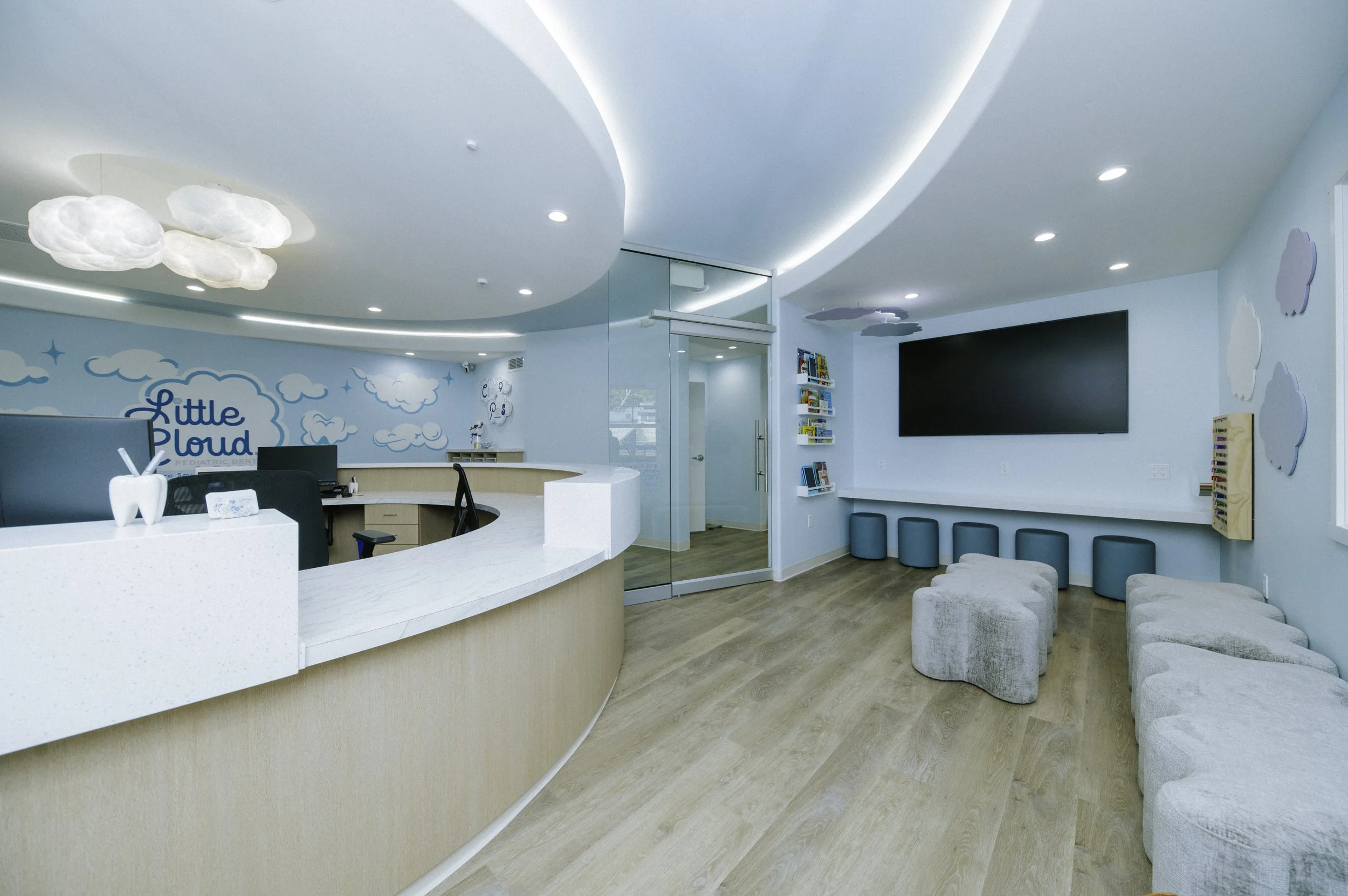 This pediatric dentist's office features curved dropped ceilings with recessed lighting that add interest and accoustics. Cloud shaped benches and kid sized ottomans offer seating in the waiting room. Cloud pendants seem to float above the front desk