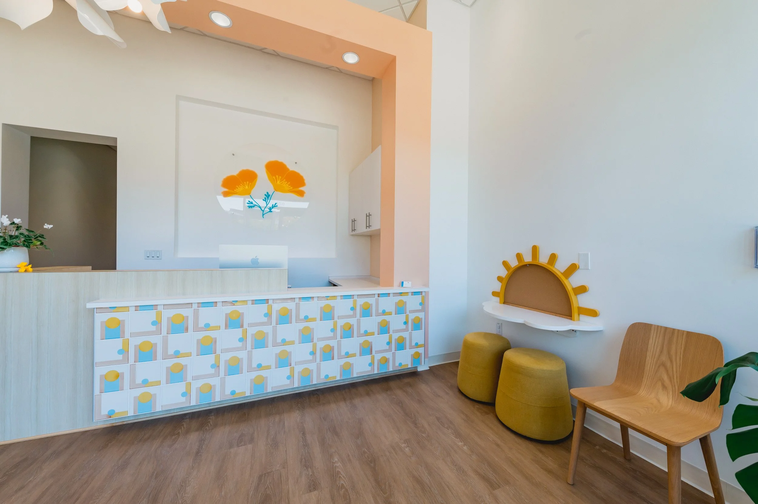 A reception area with a pastel-colored counter decorated with abstract geometric patterns, a wall art of orange flowers, two yellow stools, a wooden chair, and decorative sun motif on the wall.