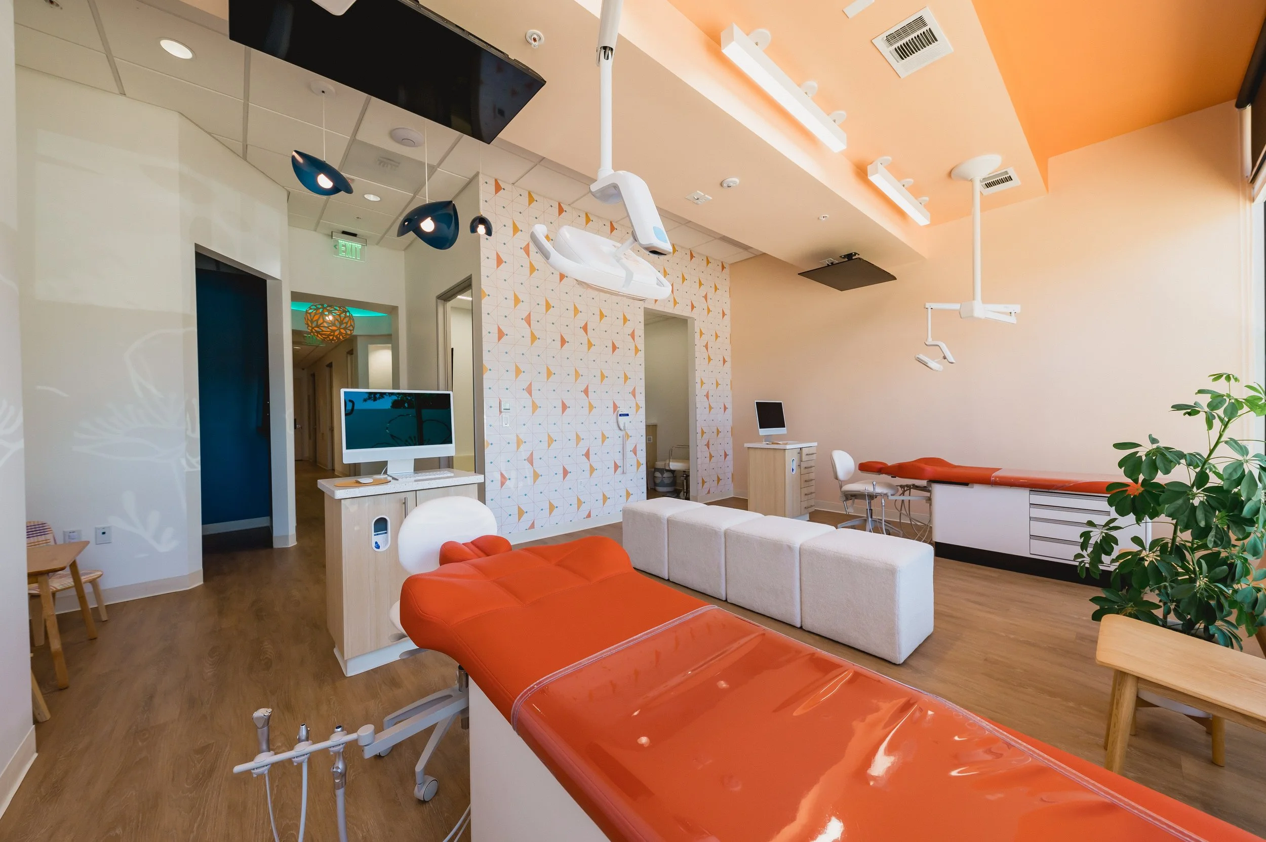 A modern dental examination room with dental chairs, monitors, and medical equipment, decorated with light-colored walls, orange accents, and wooden flooring.