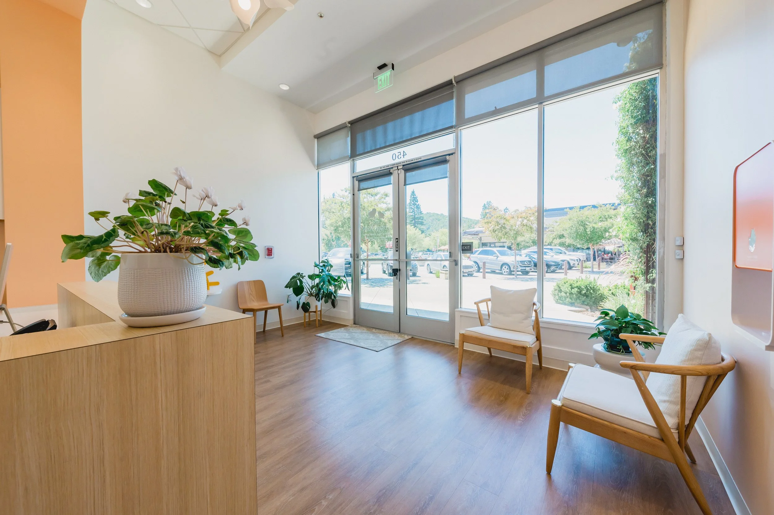 Lobby area with wooden flooring, beige walls, and large glass front door. There are two wooden chairs with cushions, potted plants, and a flower arrangement on a reception counter.