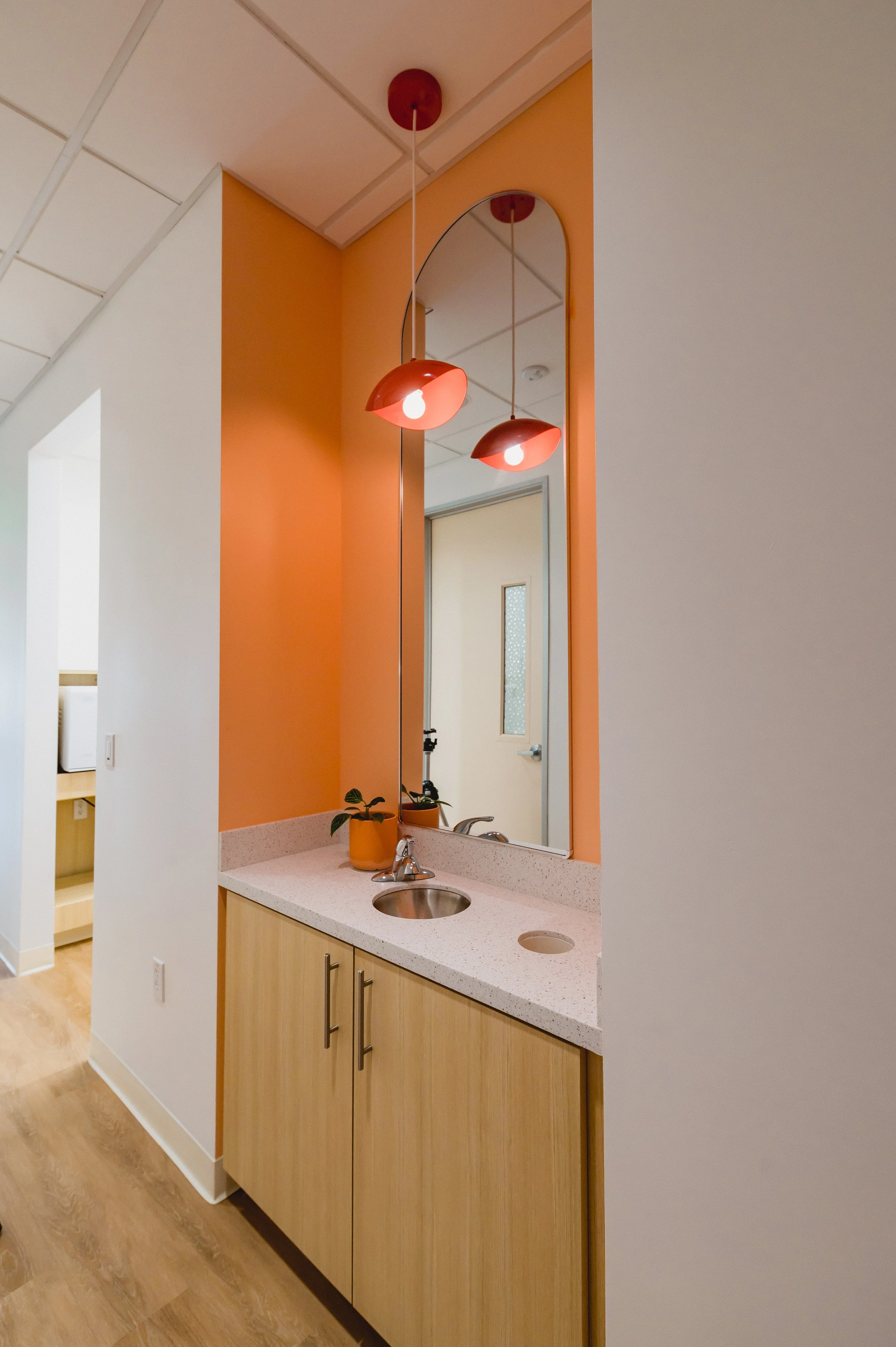 A bathroom vanity with a large mirror, minimal countertop with a sink, a small potted plant, and modern pendant lights hanging above, with an orange wall in the background.