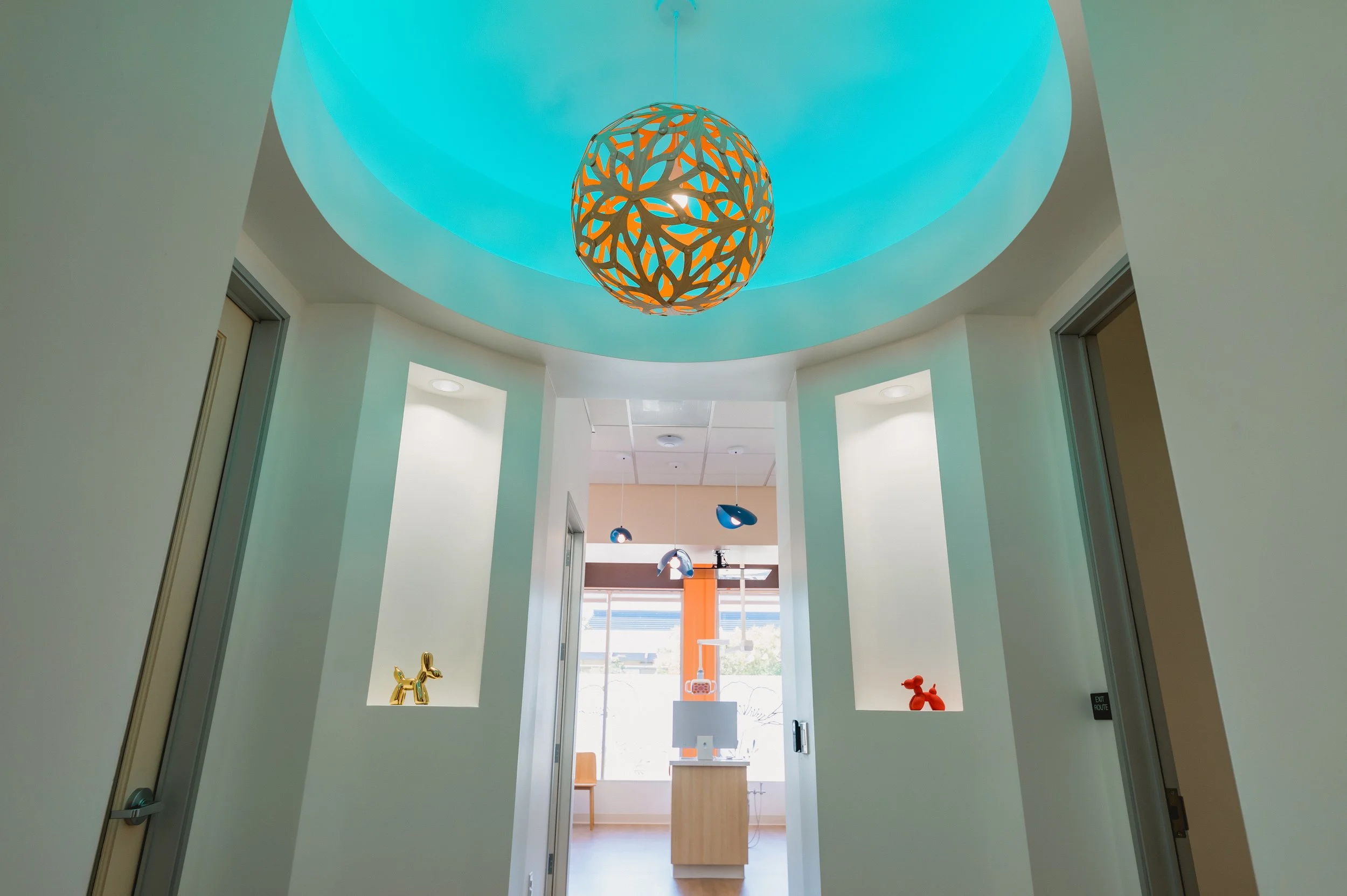 Interior view of a modern room with a blue ceiling and a decorative orange hanging light fixture. There are two shelves with small decorative items, a gold dog figure and an orange abstract sculpture. Sunlight streams in through large windows at the 