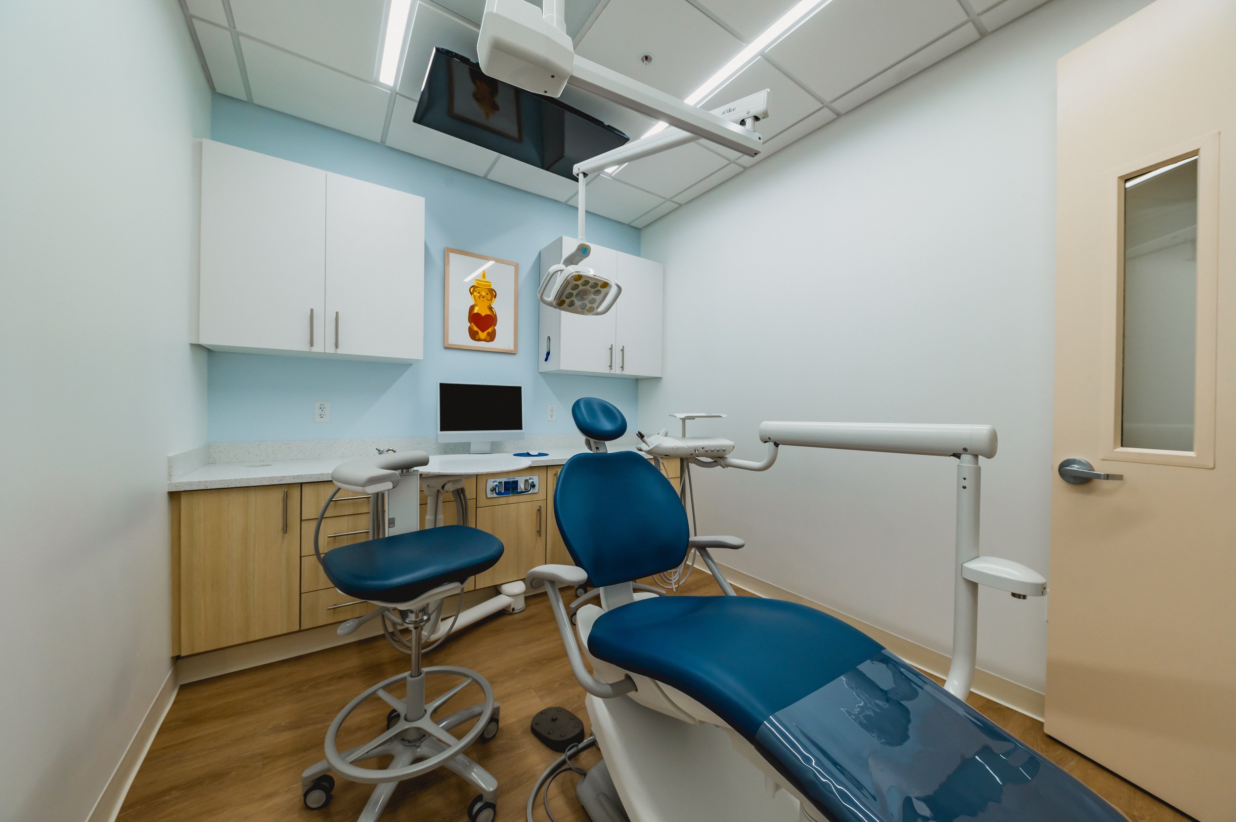 Empty dental examination room with dental chair, stool, overhead light, monitor, cabinets, and door.