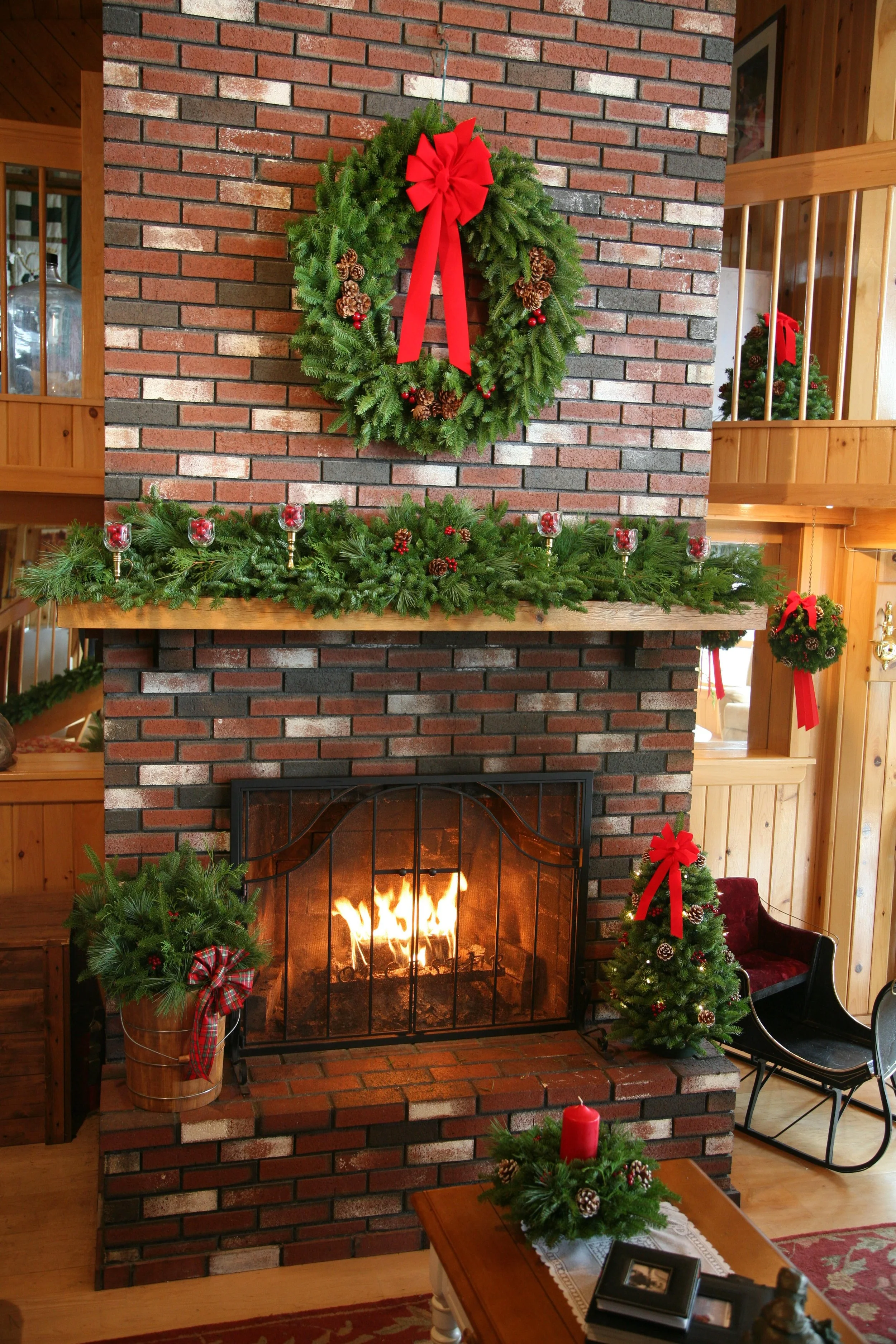 Christmas decorated brick fireplace with green wreath above, garland with candles on mantle, small decorated tree with red bow to the right, and a red candle on a table in front.