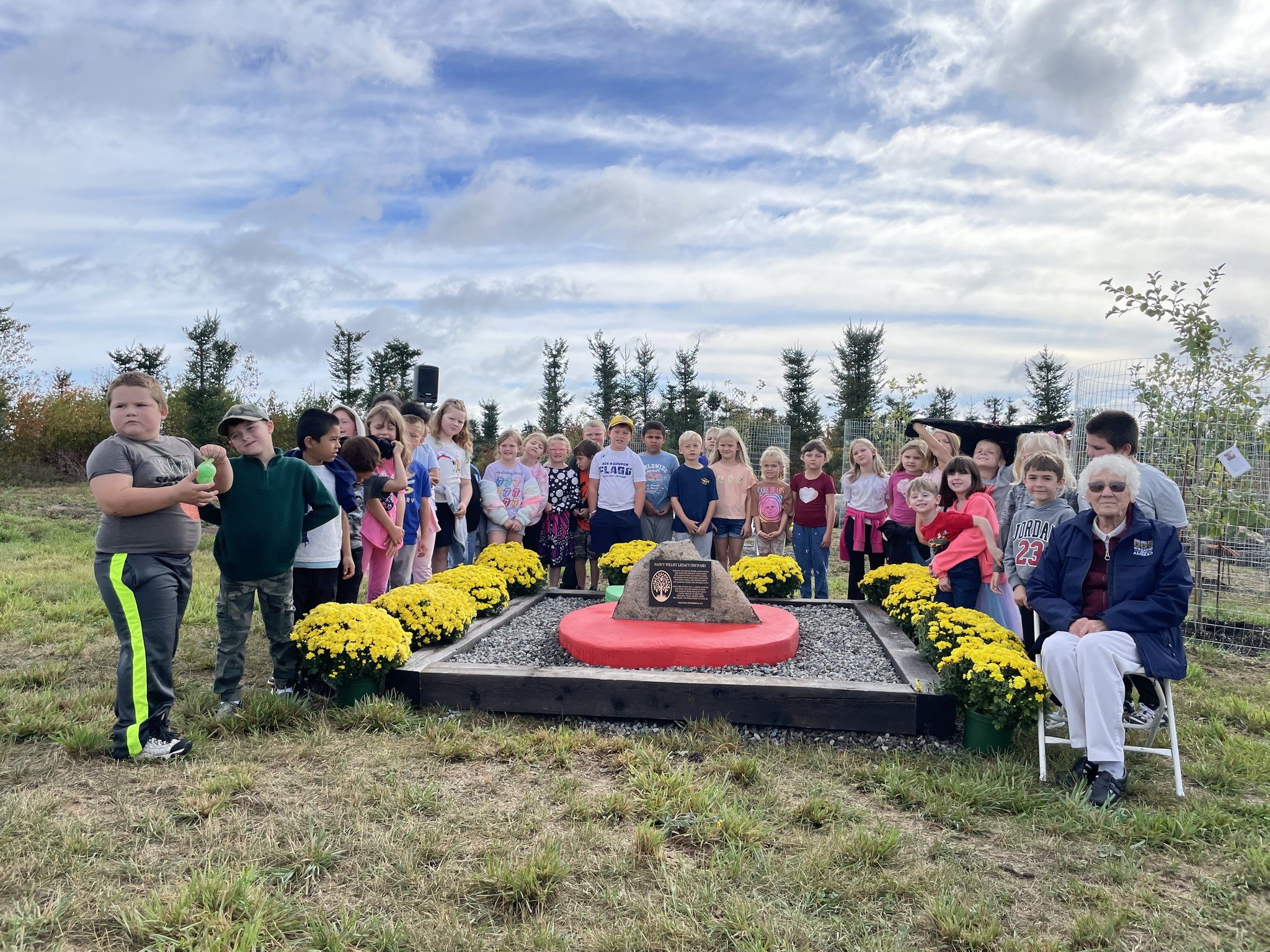 Children and an elderly woman gathered around a memorial with yellow flowers, during an outdoor ceremony on a cloudy day.