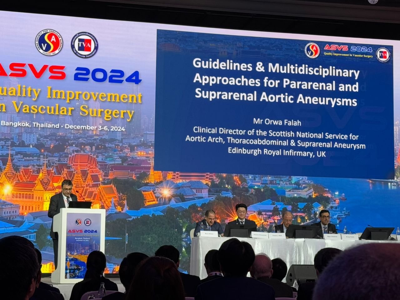 A conference in Bangkok, Thailand, with a speaker at a podium and four people seated at a panel table, discussing vascular surgery guidelines. A large screen displays the conference title, date, and speaker details.