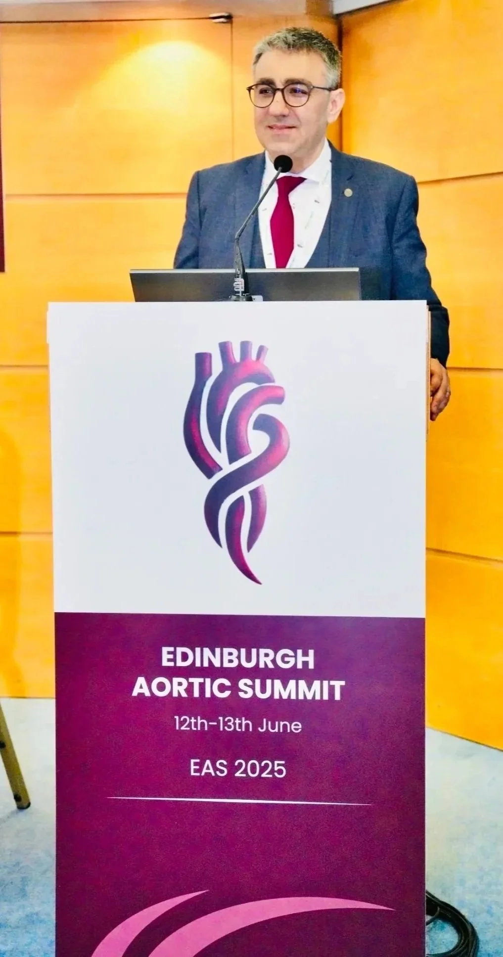A man in a suit and glasses standing behind a podium, giving a presentation at the Edinburgh Aortic Summit on June 12th-13th, 2025. The podium has a logo of a stylized heart and the event details.