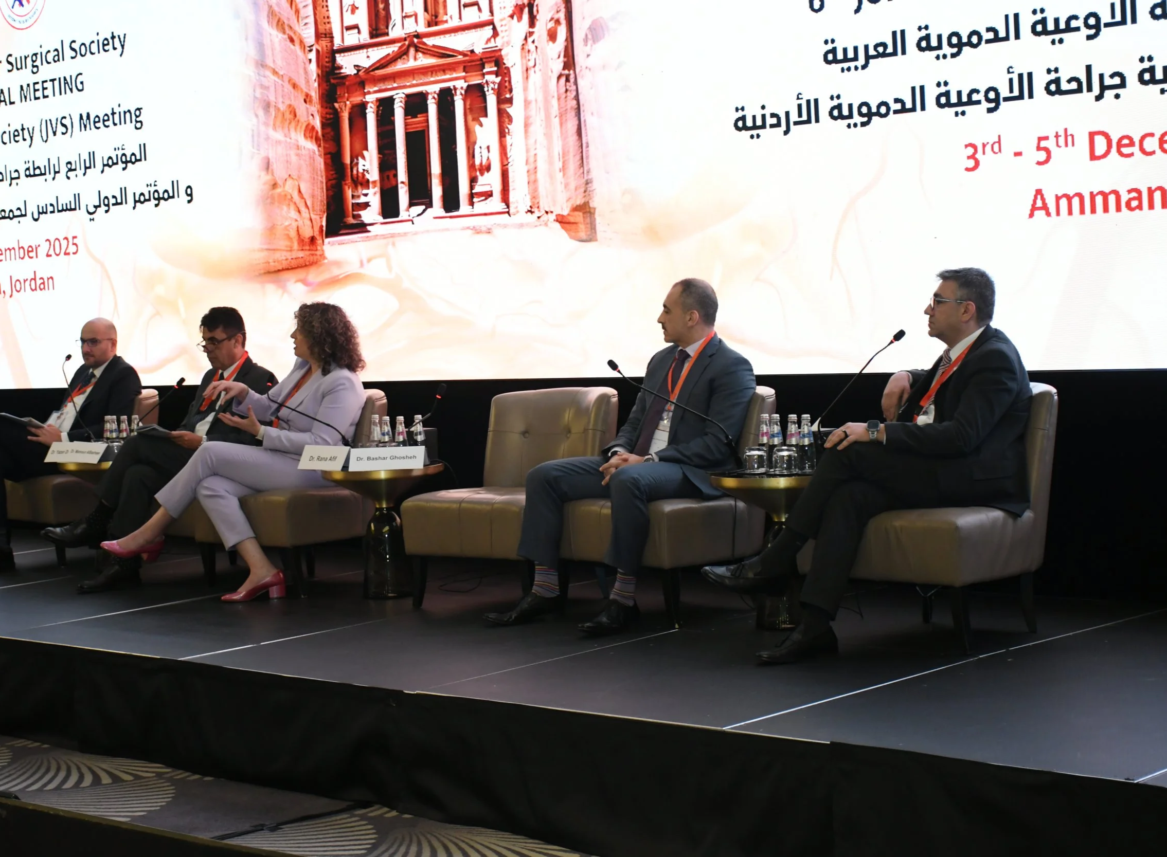 A panel of five individuals seated on stage at a conference with a large screen behind them. The screen displays text in both Arabic and English, and features an image of an ancient temple or monument. Bottles of water and glasses are placed on small tables in front of them.