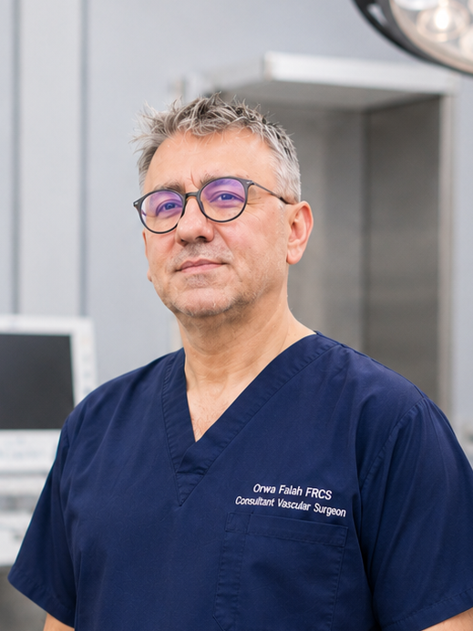 A middle-aged man with gray hair and glasses, wearing navy medical scrubs with his name and title embroidered on the chest, standing in a hospital or surgical room.