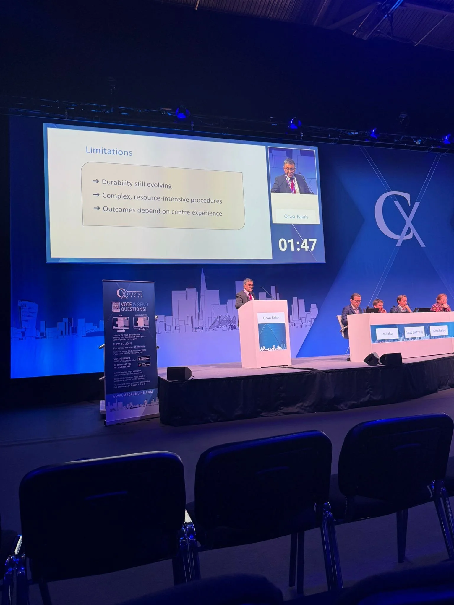 A conference or panel discussion with a large screen displaying a presentation slide titled 'Limitations' and points about durability, resource-intensive procedures, and outcomes depending on center experience. The slide includes a small video feed of a speaker, Orwa Falah, and a timer showing 01:47. Several seated panelists are on stage, and audience chairs are visible in the foreground.