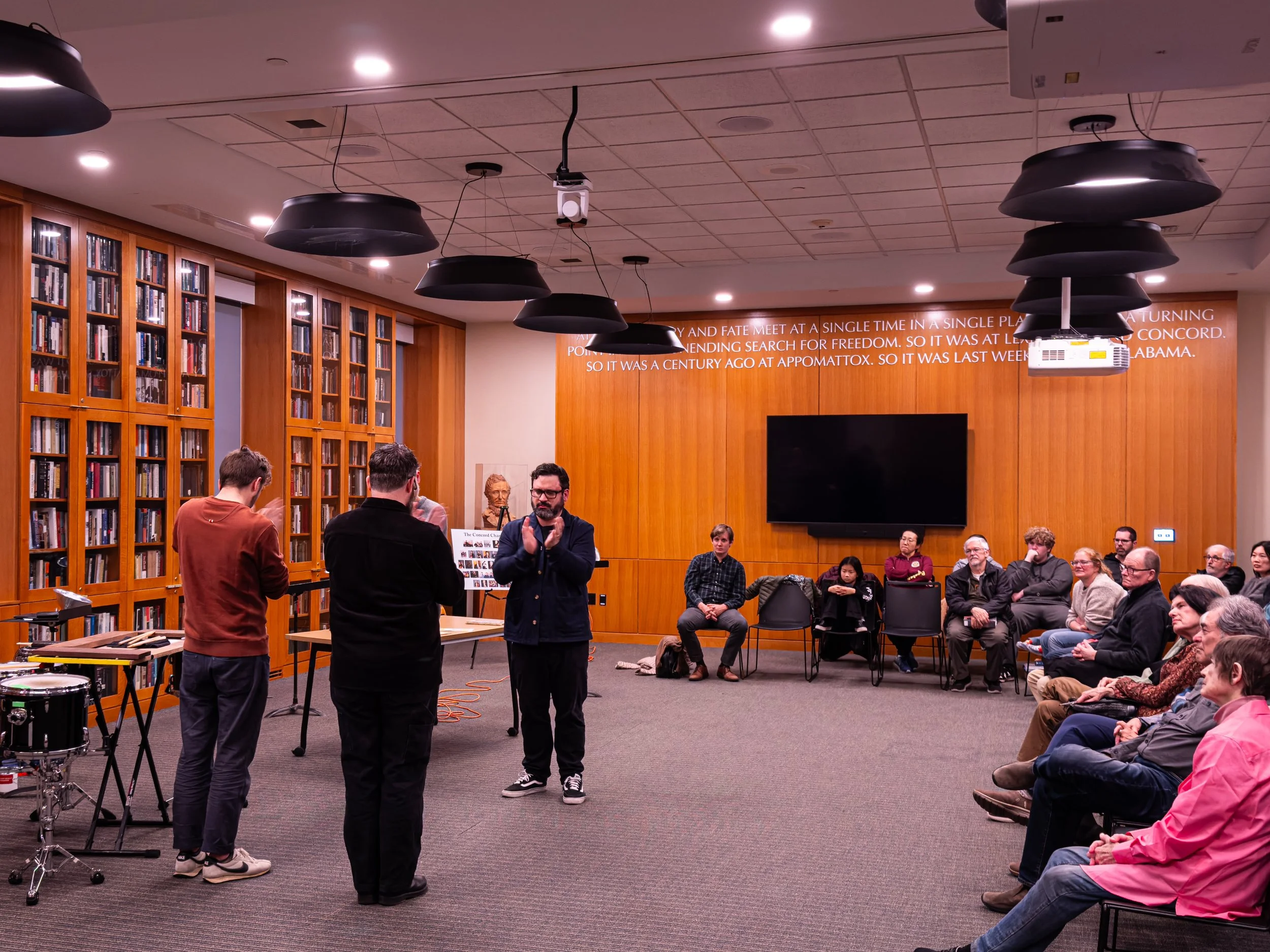 A group of people attending a presentation or panel discussion in a room with wooden paneling and built-in bookshelves filled with books. Some individuals are seated and listening, while others are standing and speaking or presenting at the front. A man is clapping, and a woman behind him is gesturing. There is a large television screen and a projector mounted on the ceiling, along with a wall display with white text on an orange background.
