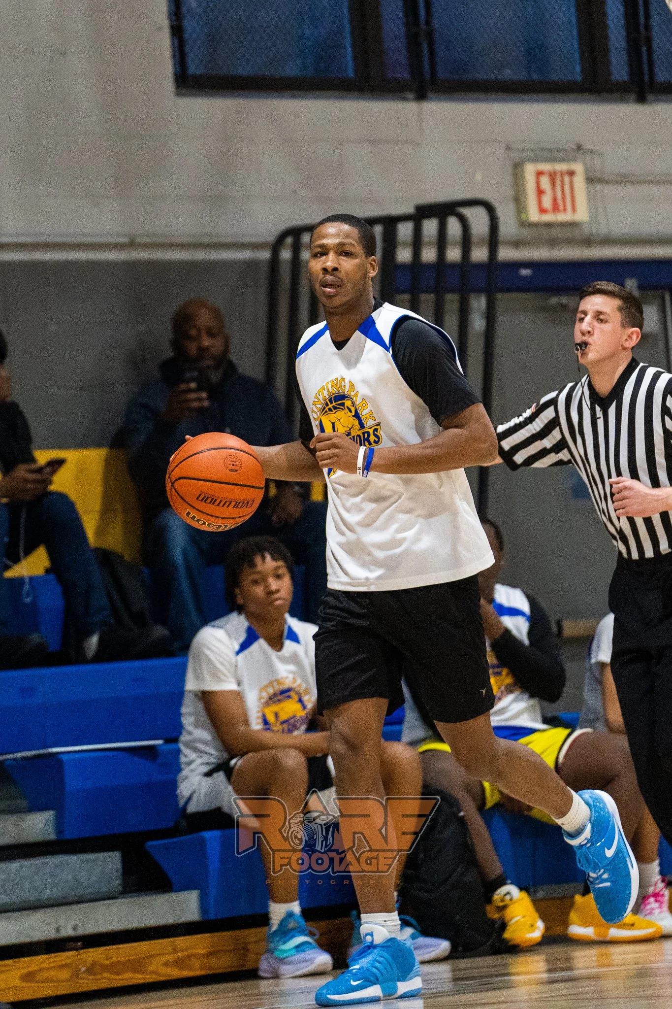 A male with black, white, and yellow shirt, bouncing a ball during a basketball game. 
