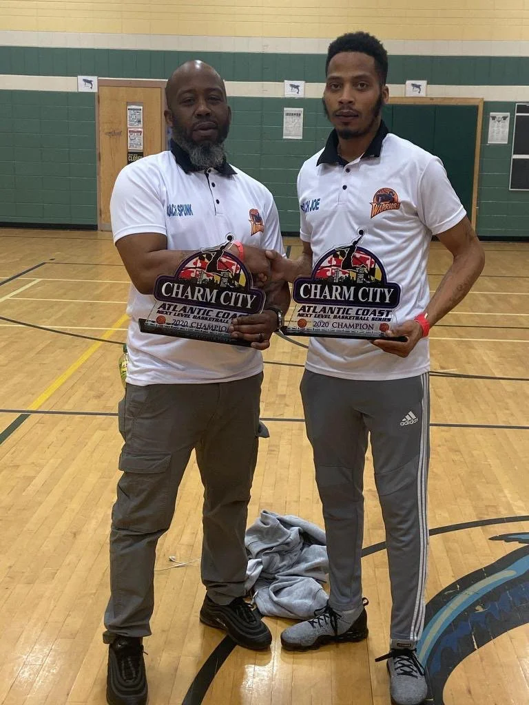 Two black males, wearing white and holding a charm city plate. 
