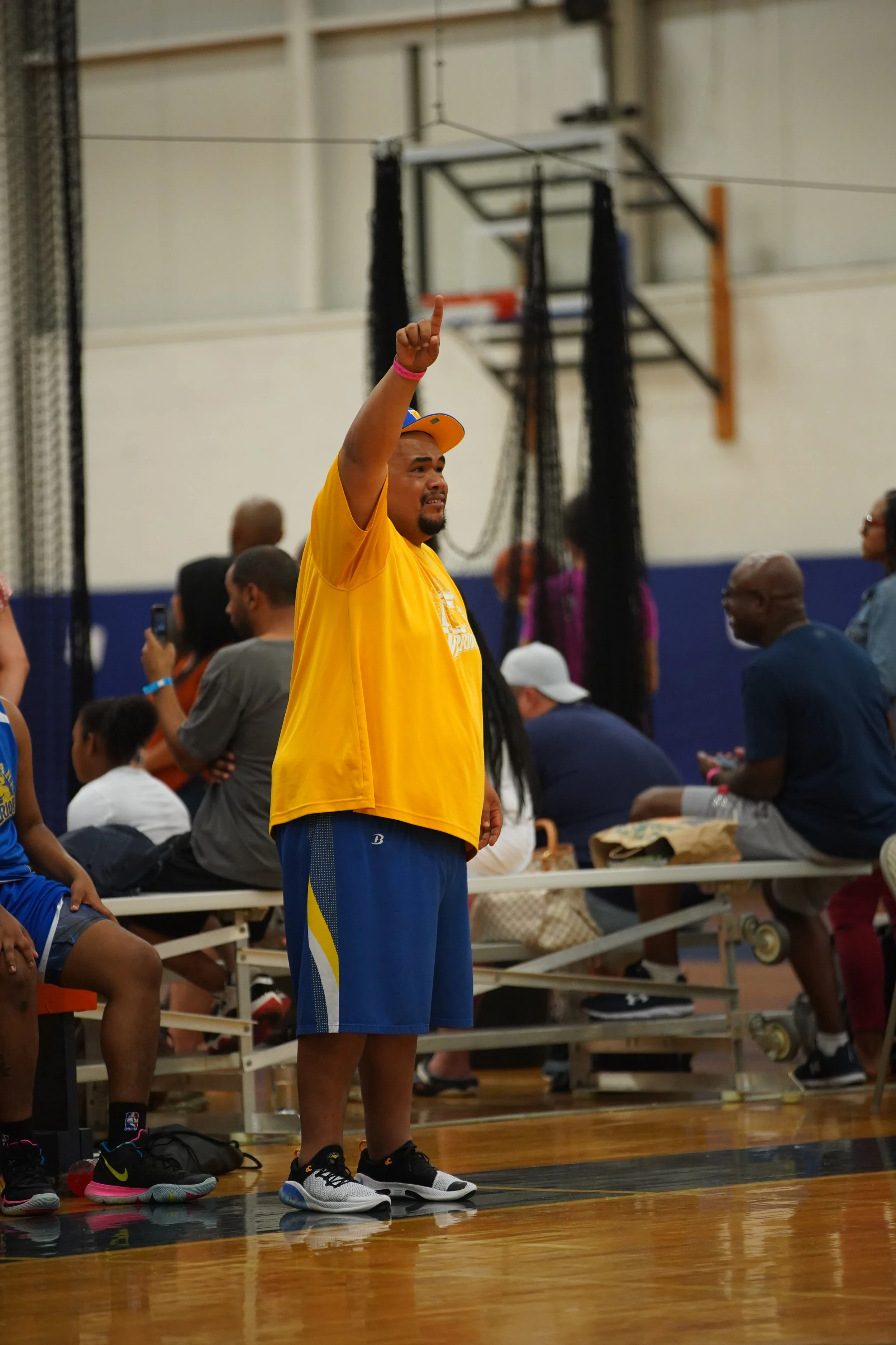 A basketball coach wearing a hat, with a yellow shirt that has a white logo, and blue shorts. 