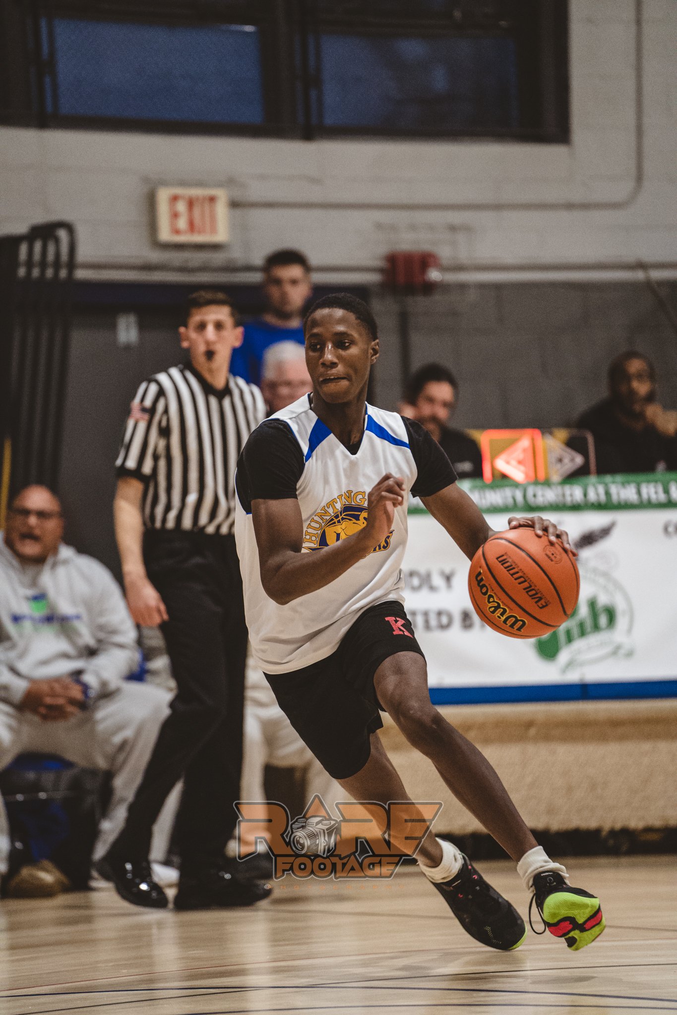 A young black male, bouncing the ball in a basketball game. Wearing black, white and yellow. 