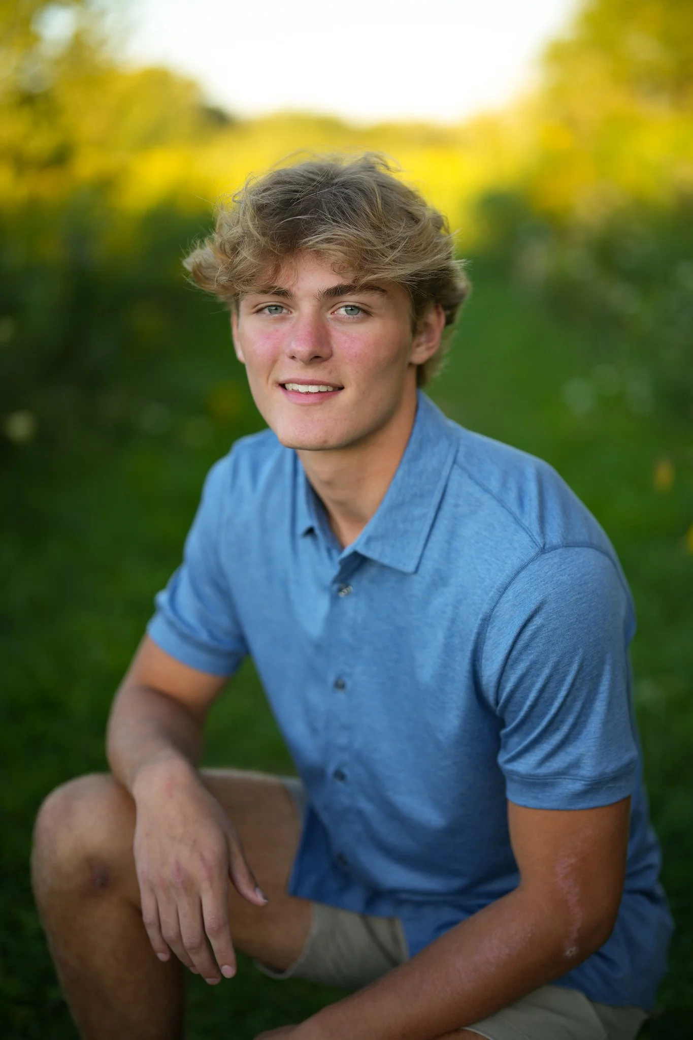 A young man with blonde hair and blue eyes wearing a blue shirt, sitting outdoors on green grass with a blurred natural background, smiling slightly at the camera.