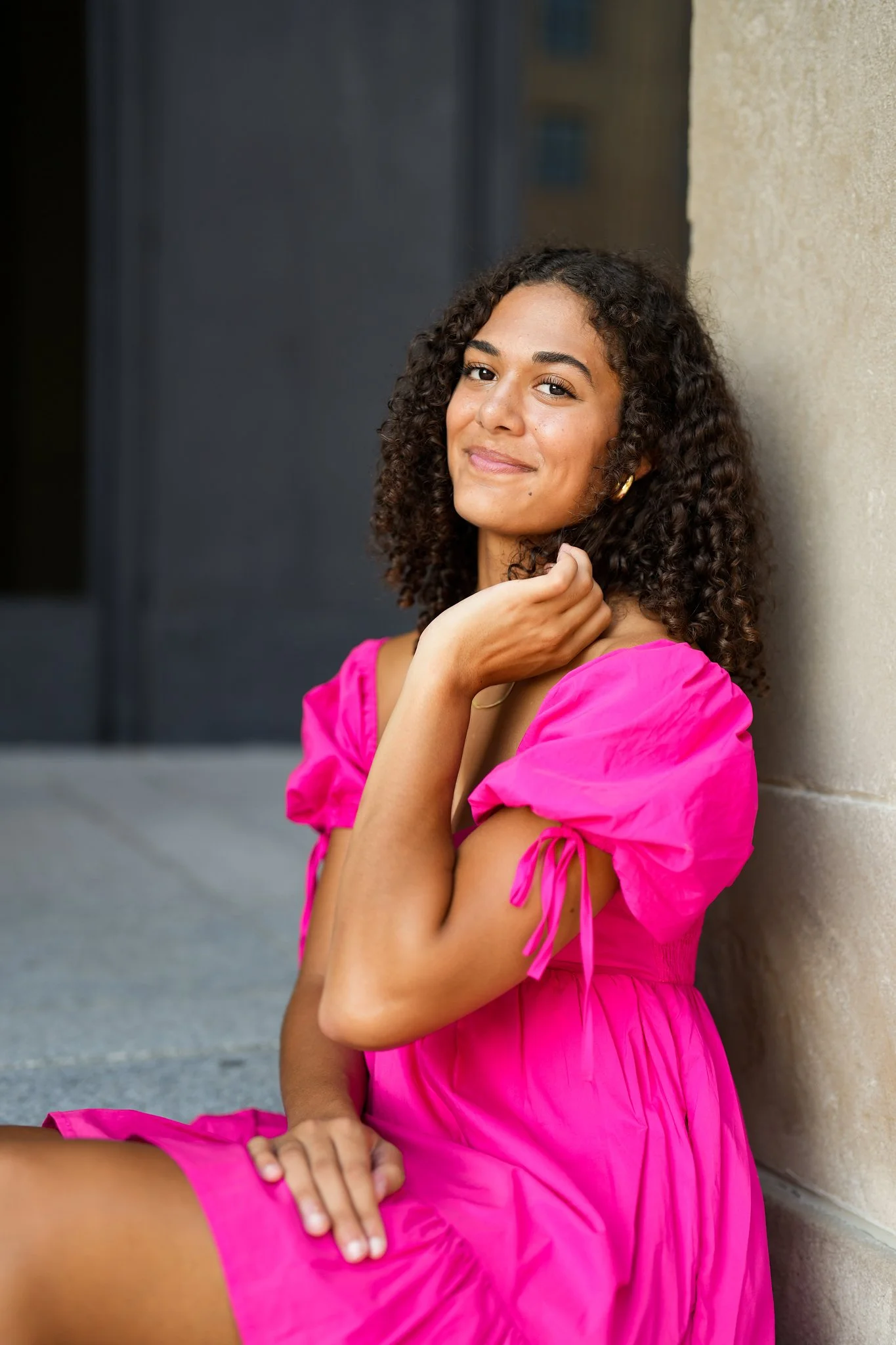A young woman with curly hair and hoop earrings, wearing a bright pink dress, sitting against a wall, smiling at the camera.