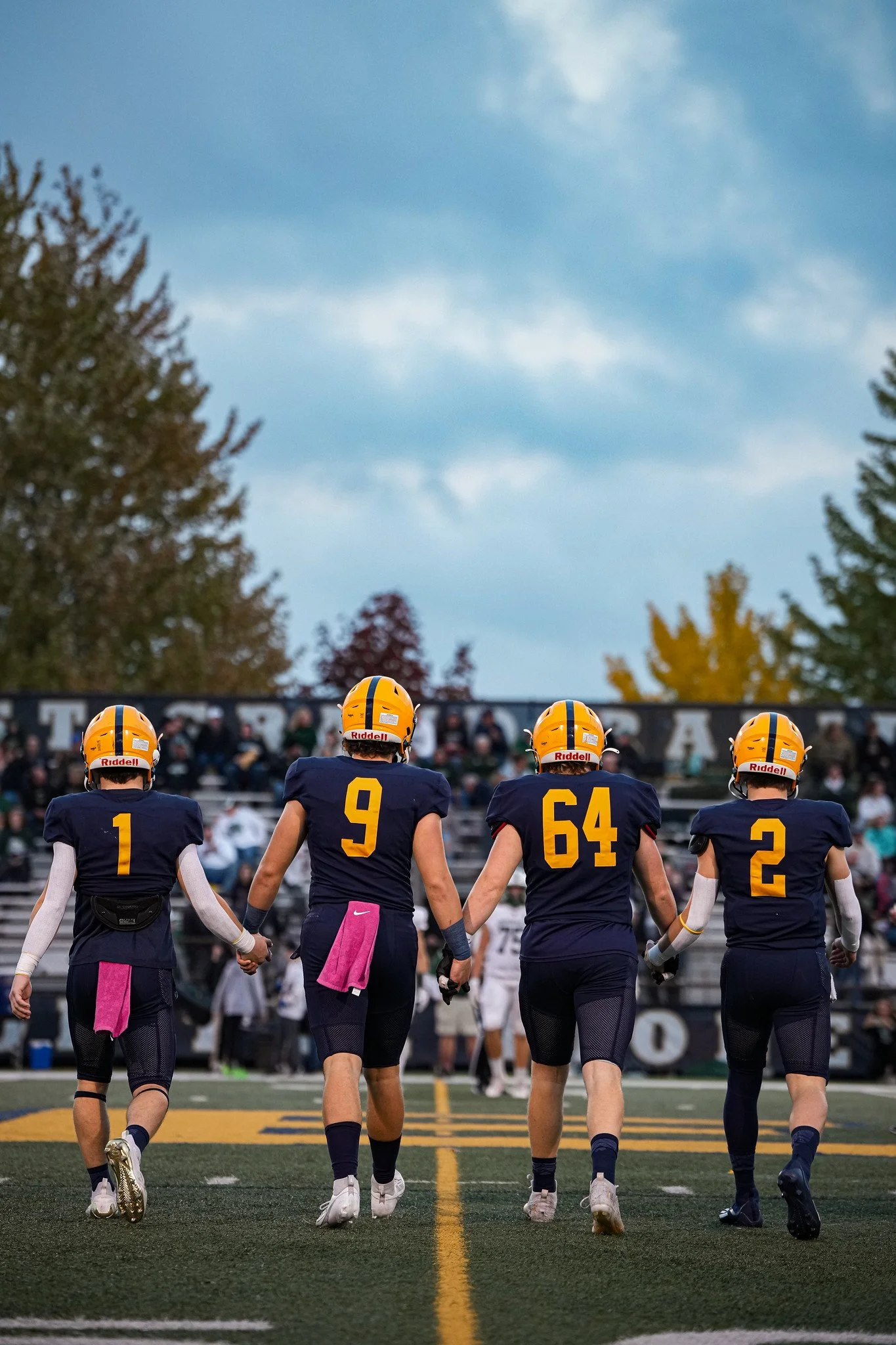 Football players walking hand in hand on the field, wearing blue uniforms with yellow helmets, numbered 1, 9, 54, and 2, during a game at dusk with an audience in the background.