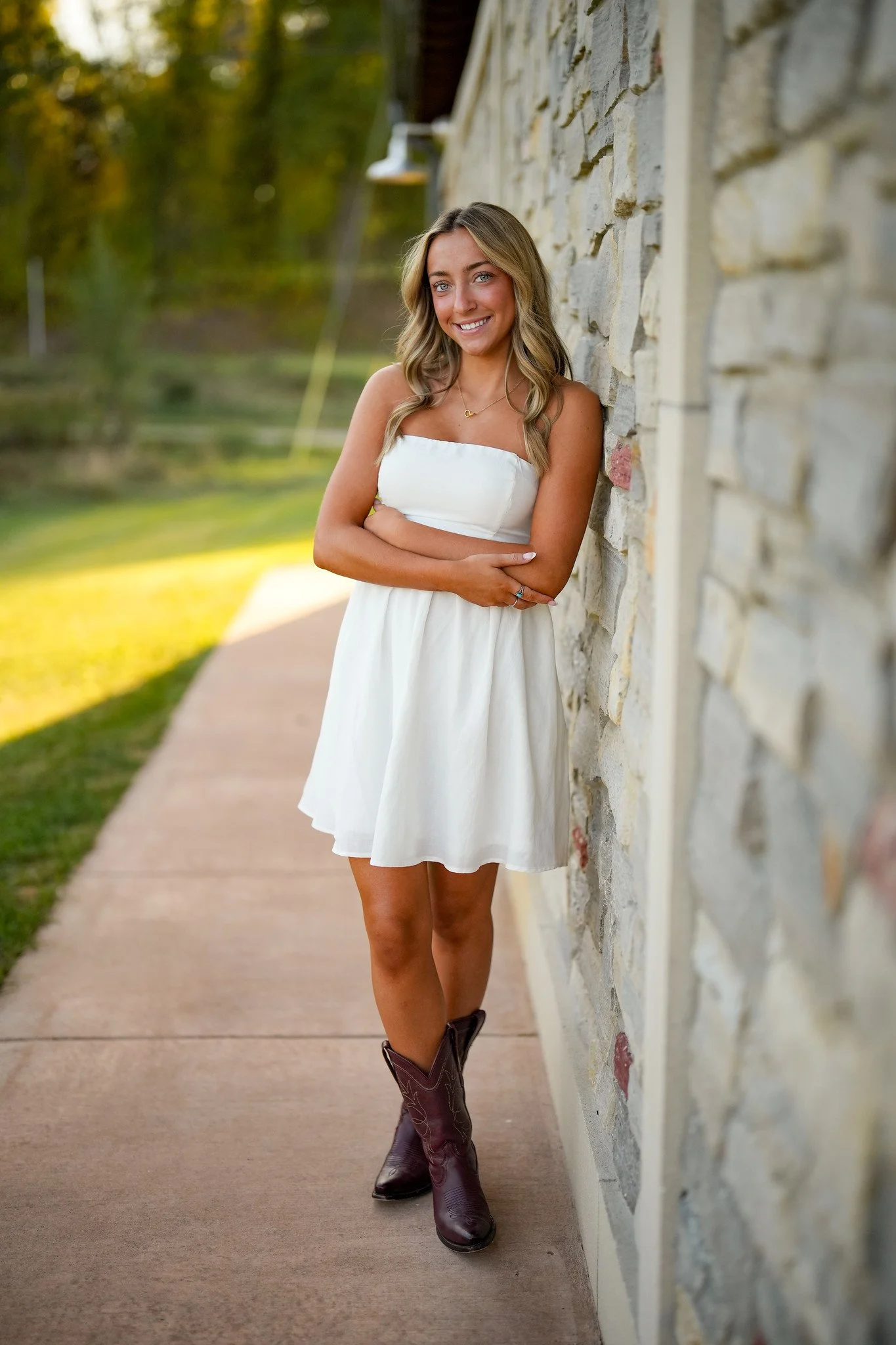 A young woman with wavy blonde hair wearing a white strapless dress, brown cowboy boots, and a necklace, leaning against a stone wall outdoors with trees and grass in the background.