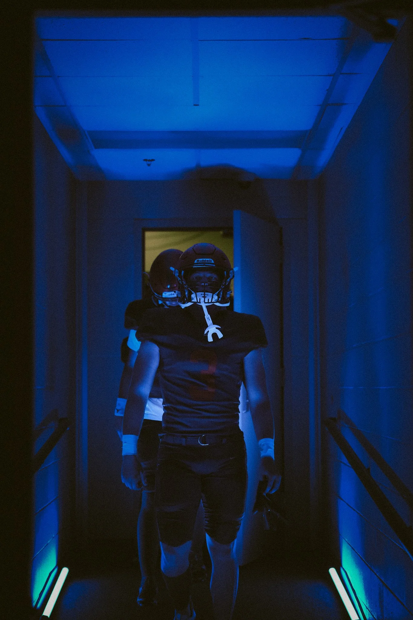 Group of football players walking through a tunnel with blue lighting, wearing helmets and sportswear.