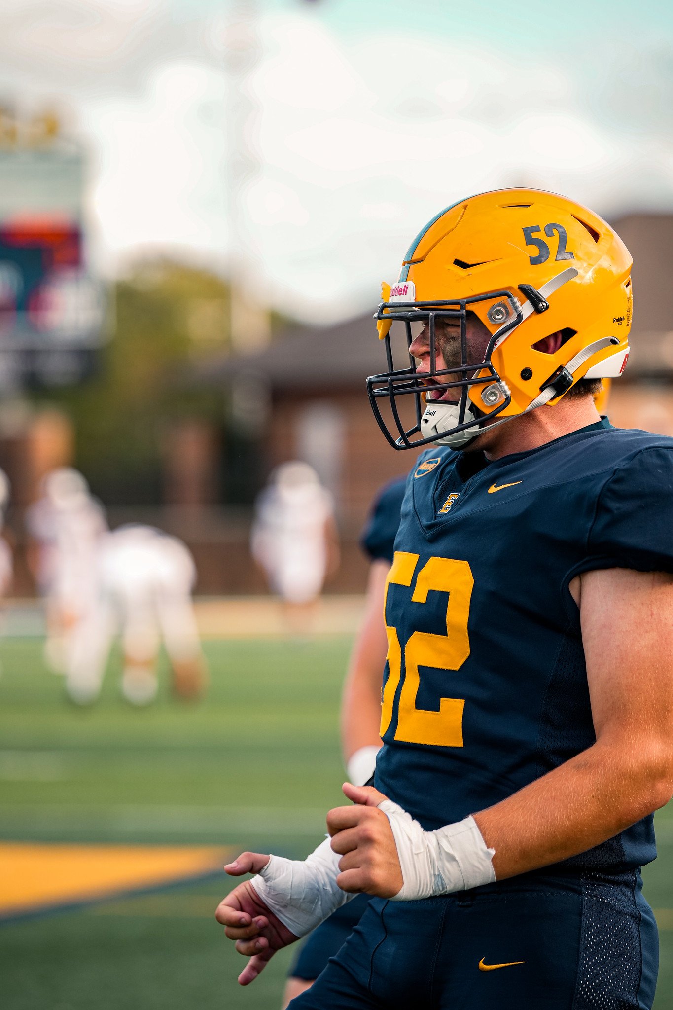 A football player wearing a yellow helmet and navy blue uniform with the number 52 is standing on a field, with other players in the background.