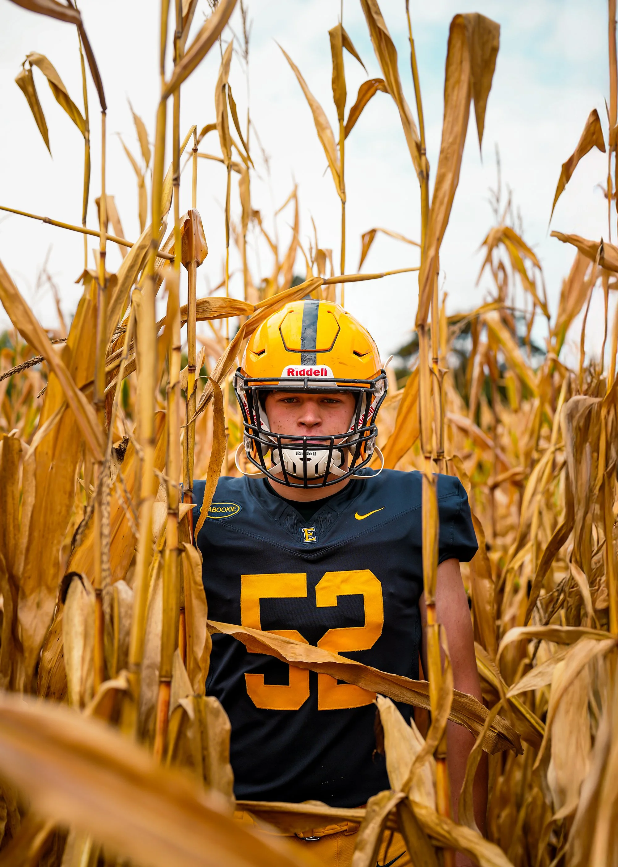 Young football player wearing a yellow helmet and a navy blue jersey with the number 52, standing in a cornfield.