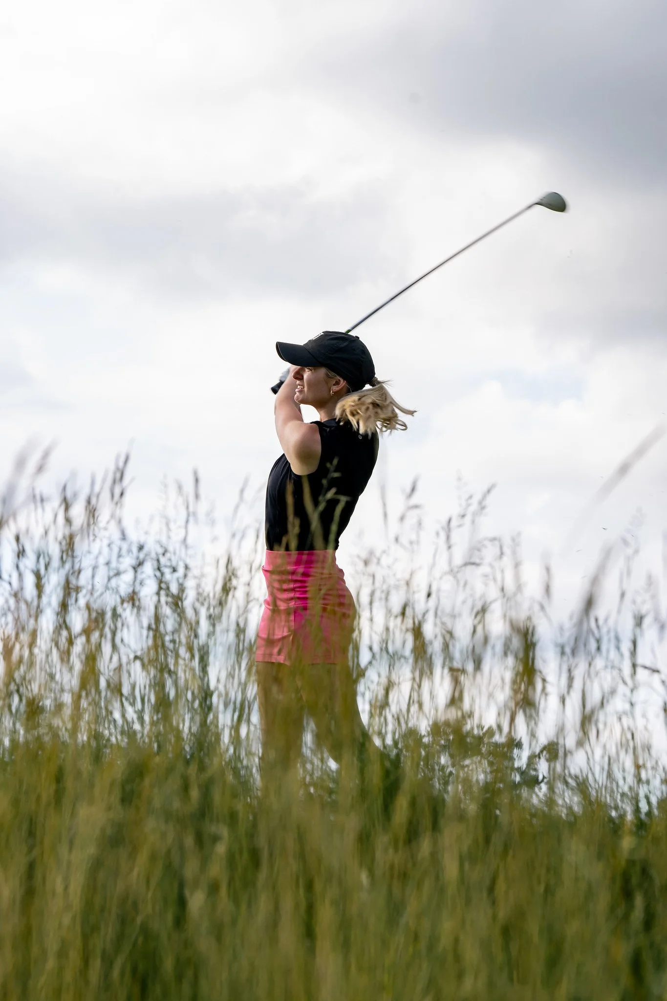 A woman in a black cap and black top with pink shorts is standing in a field of tall grass under a cloudy sky, swinging a golf club.
