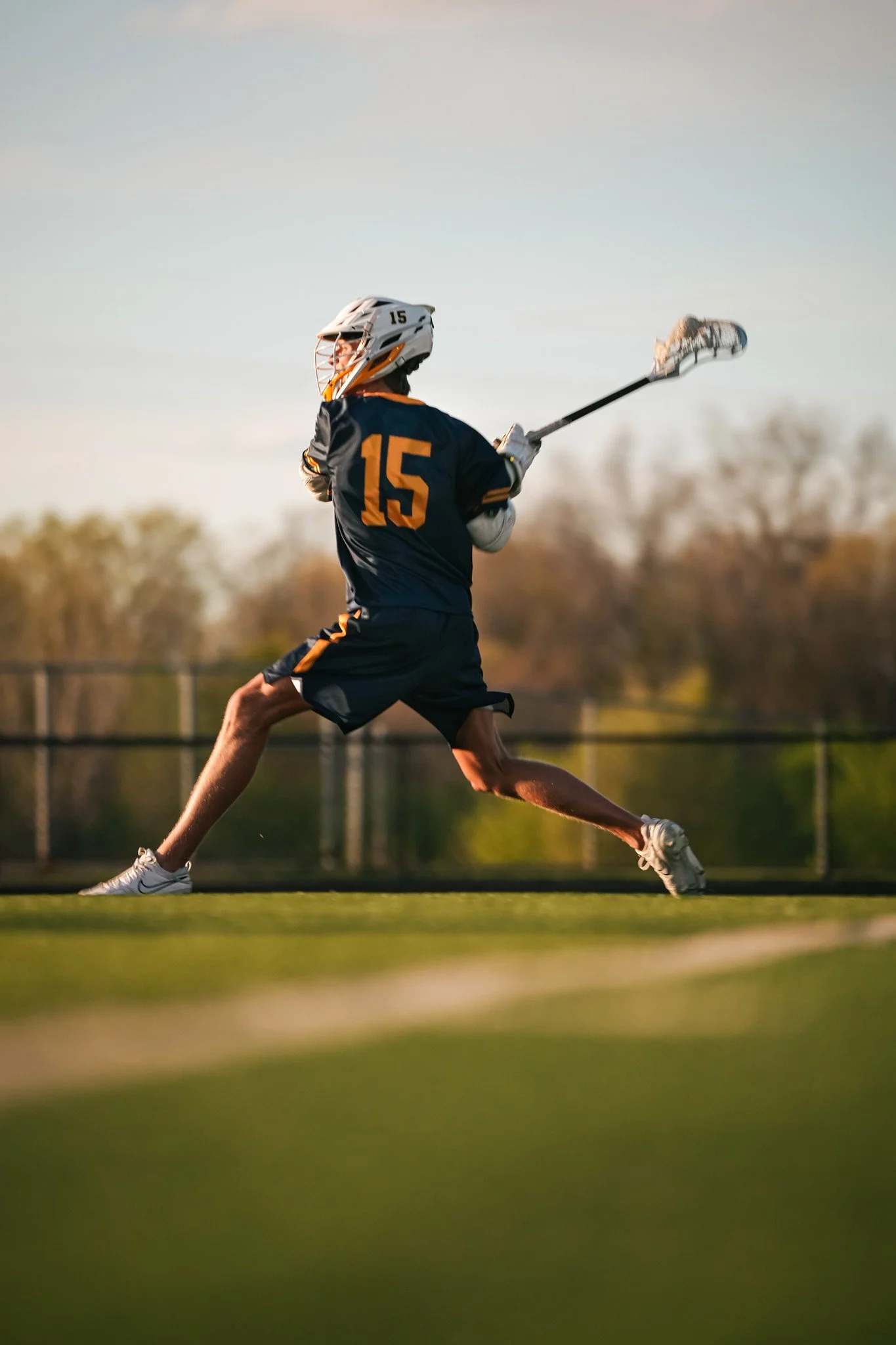 A lacrosse player wearing a navy blue and yellow uniform with the number 15 is running across the field holding a lacrosse stick in an outdoor setting during sunset.