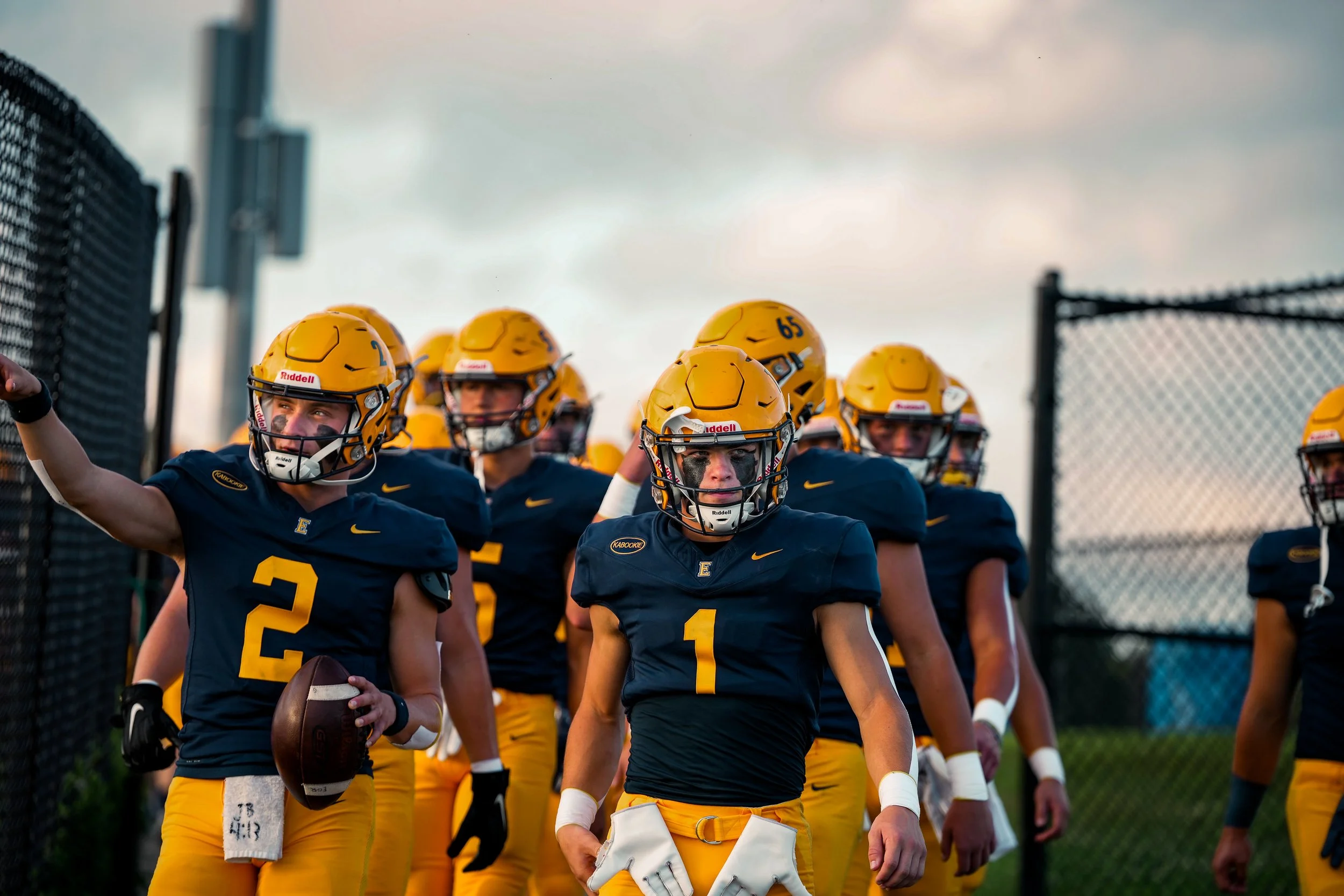 A group of football players in blue and yellow uniforms, wearing yellow helmets with face masks, standing on a football field during a game or practice. One player in the front holds a football, and others are gathered behind him, some with their arm