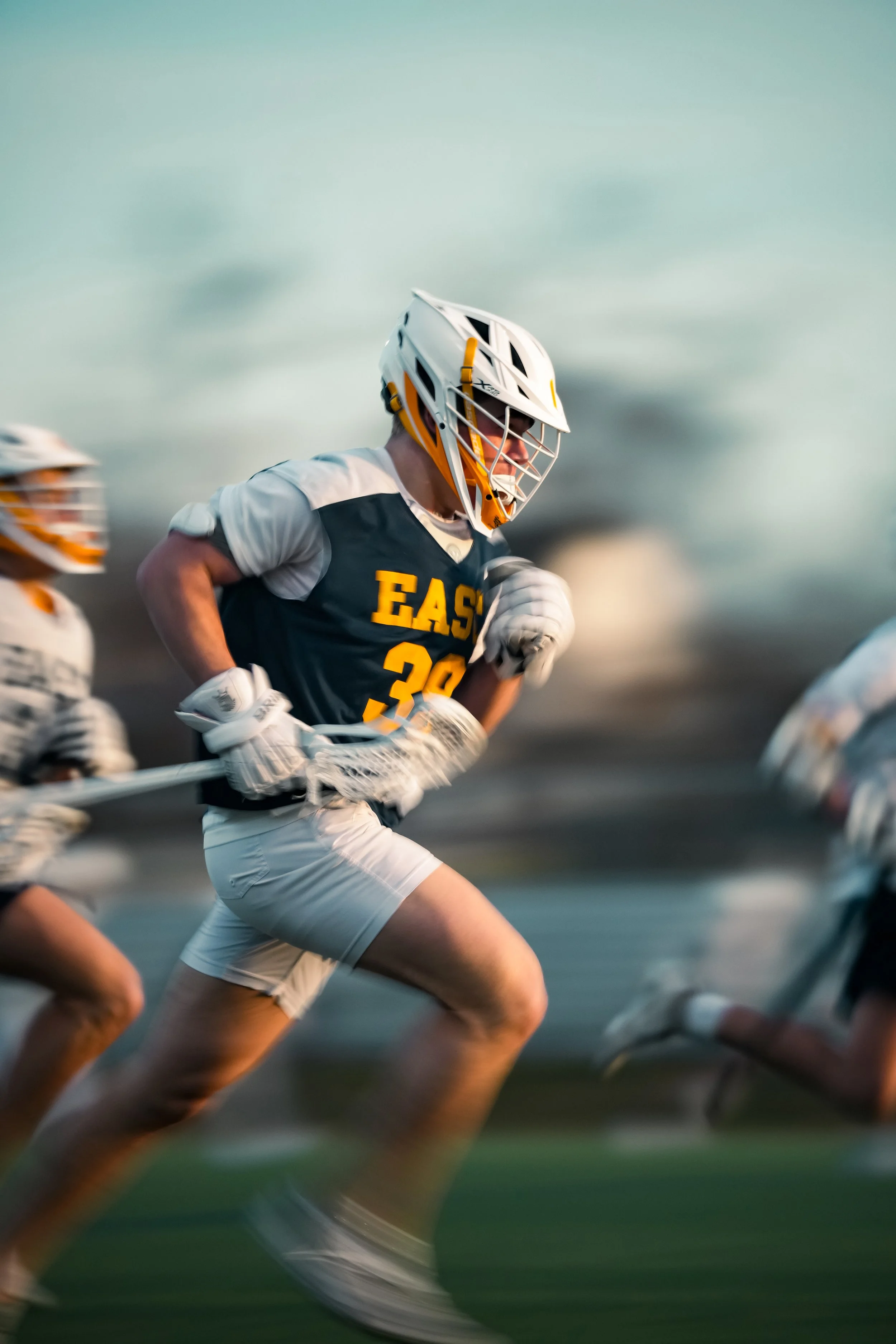 A group of lacrosse players running on the field with a cloudy sky in the background.