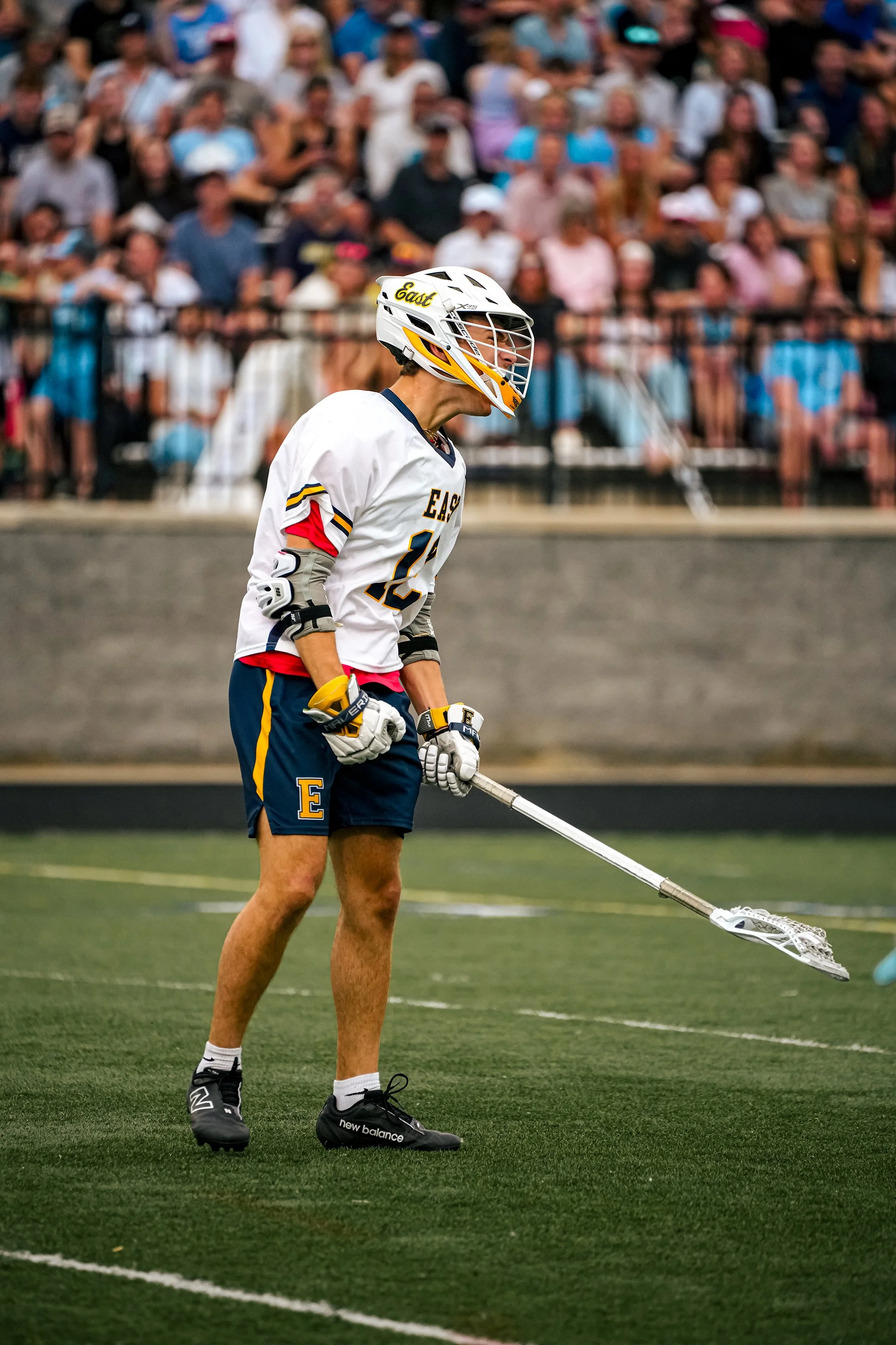A lacrosse player in a white jersey with the word 'East' is preparing to play on a green turf field, with a crowd of spectators in the background.