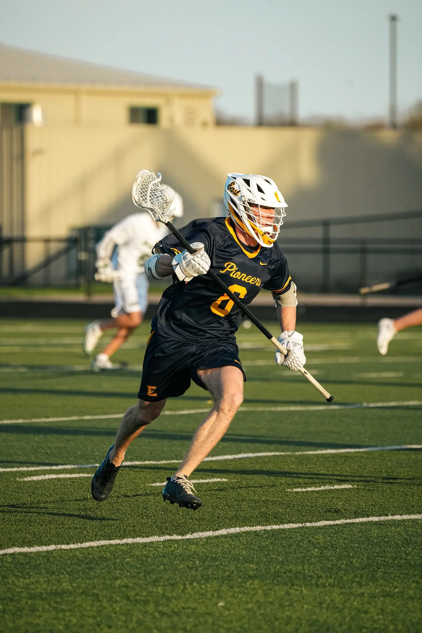 A male lacrosse player wearing a black jersey with yellow accents and the word 'Pioneers' on it, running on a green turf field with a lacrosse stick in hand. There is a person in a white jersey in the background and a building with a fence behind, wi
