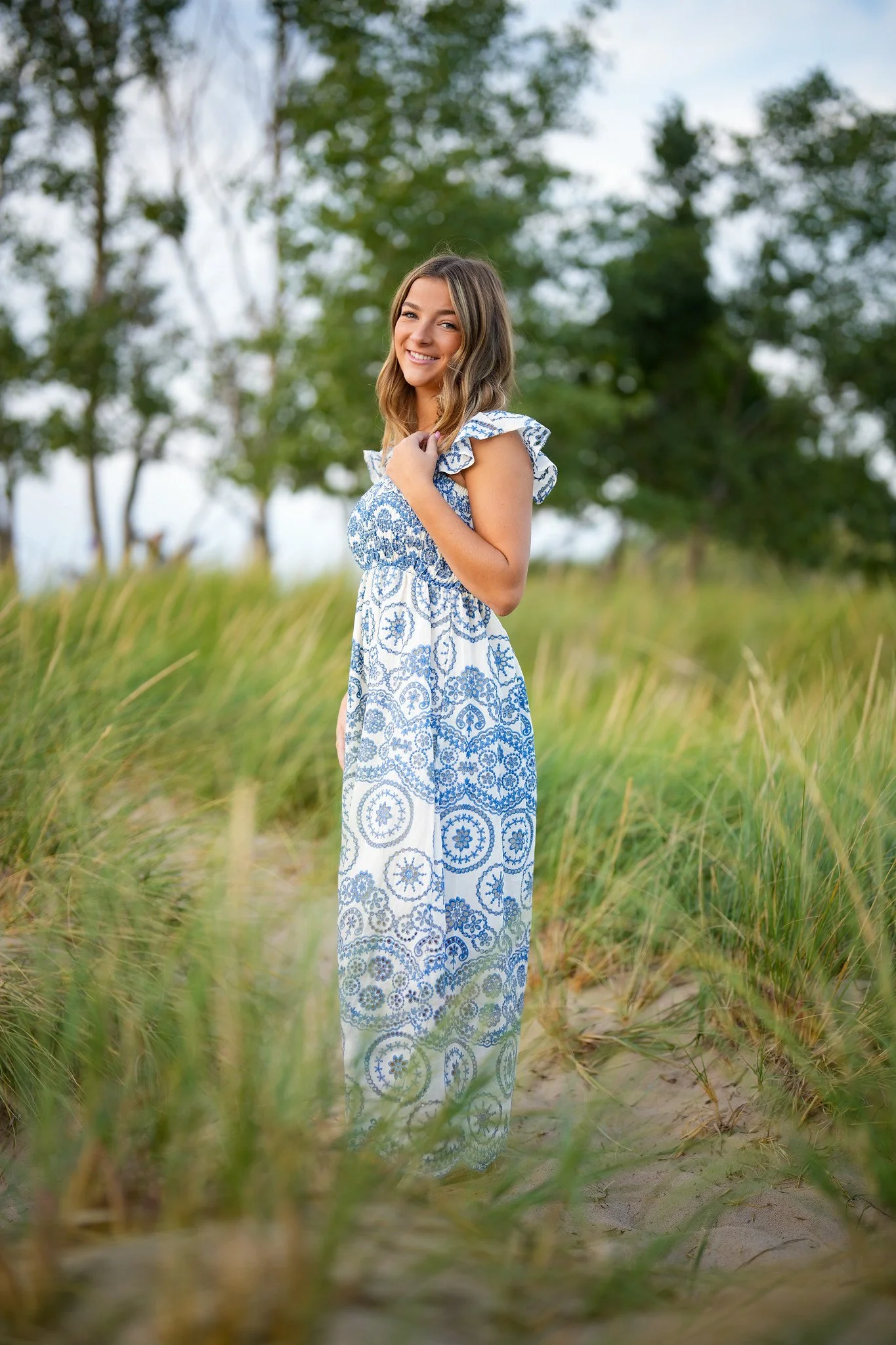 A young woman with long wavy hair wearing a blue and white patterned sundress, smiling and standing in a grassy outdoor area during daytime with trees in the background.