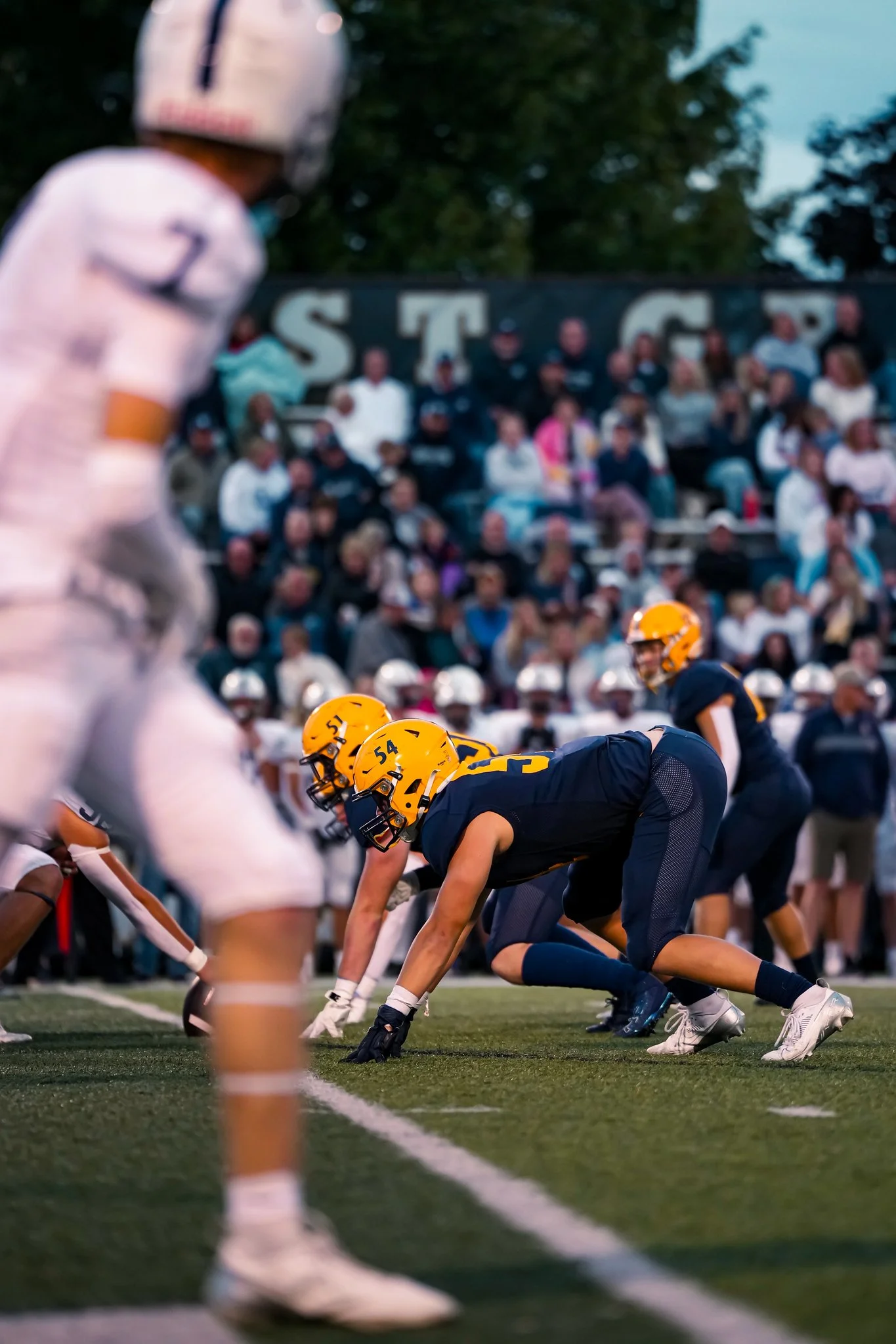 A football game at sunset with players on the field and spectators in the stands.