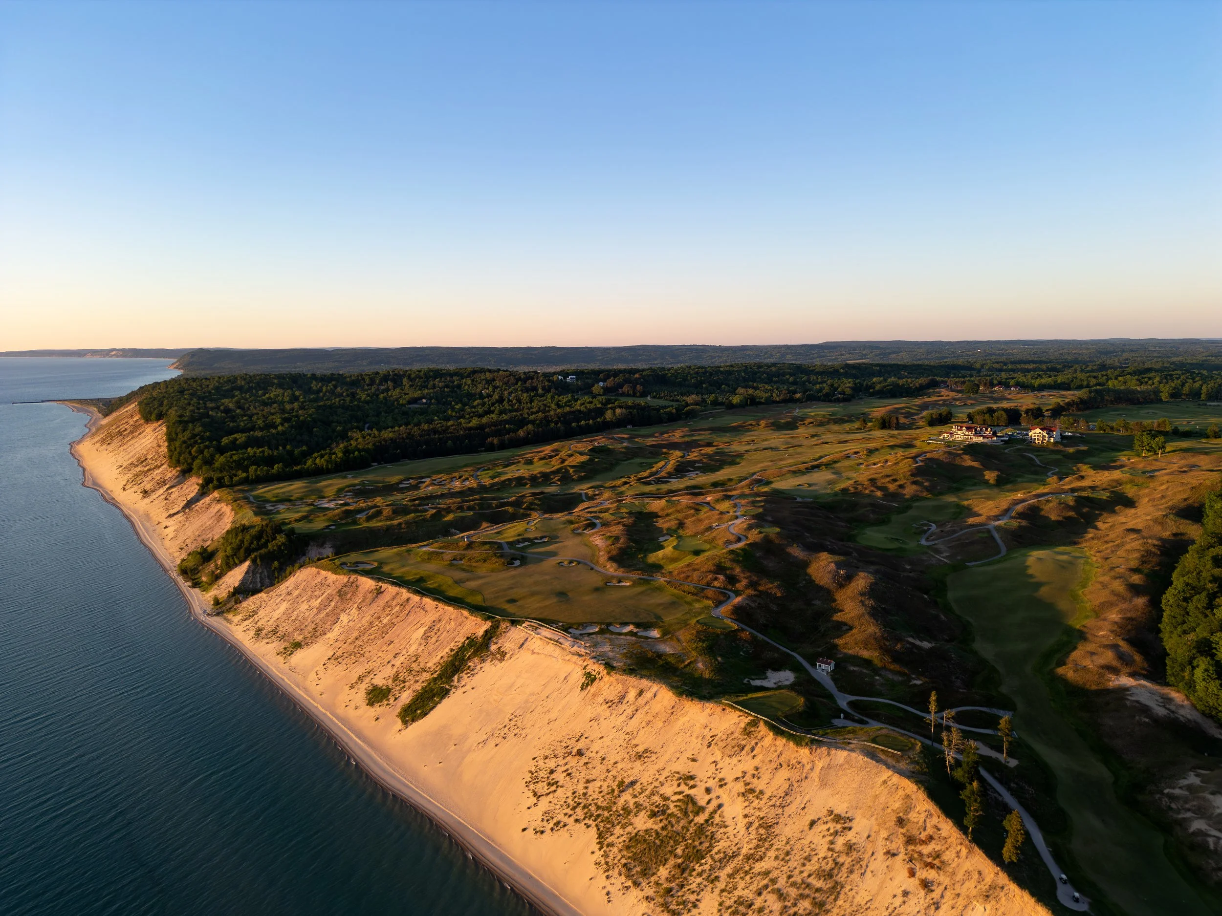 Aerial view of a coastline with a sandy beach, green hills, and a golf course at sunset.