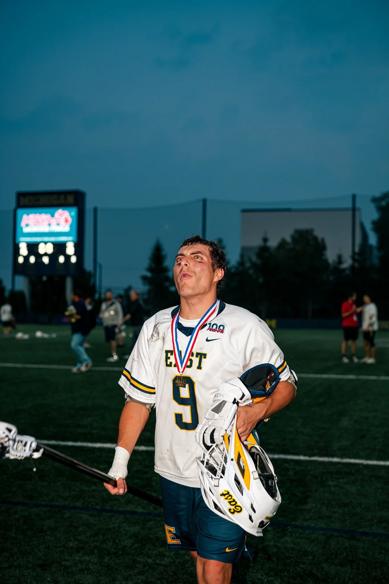 A male athlete in a white and yellow sports uniform with the number 9, holding a hockey stick and a white Goalie helmet, standing on a sports field after a game. He has a medal around his neck and is surrounded by other players and spectators in the 