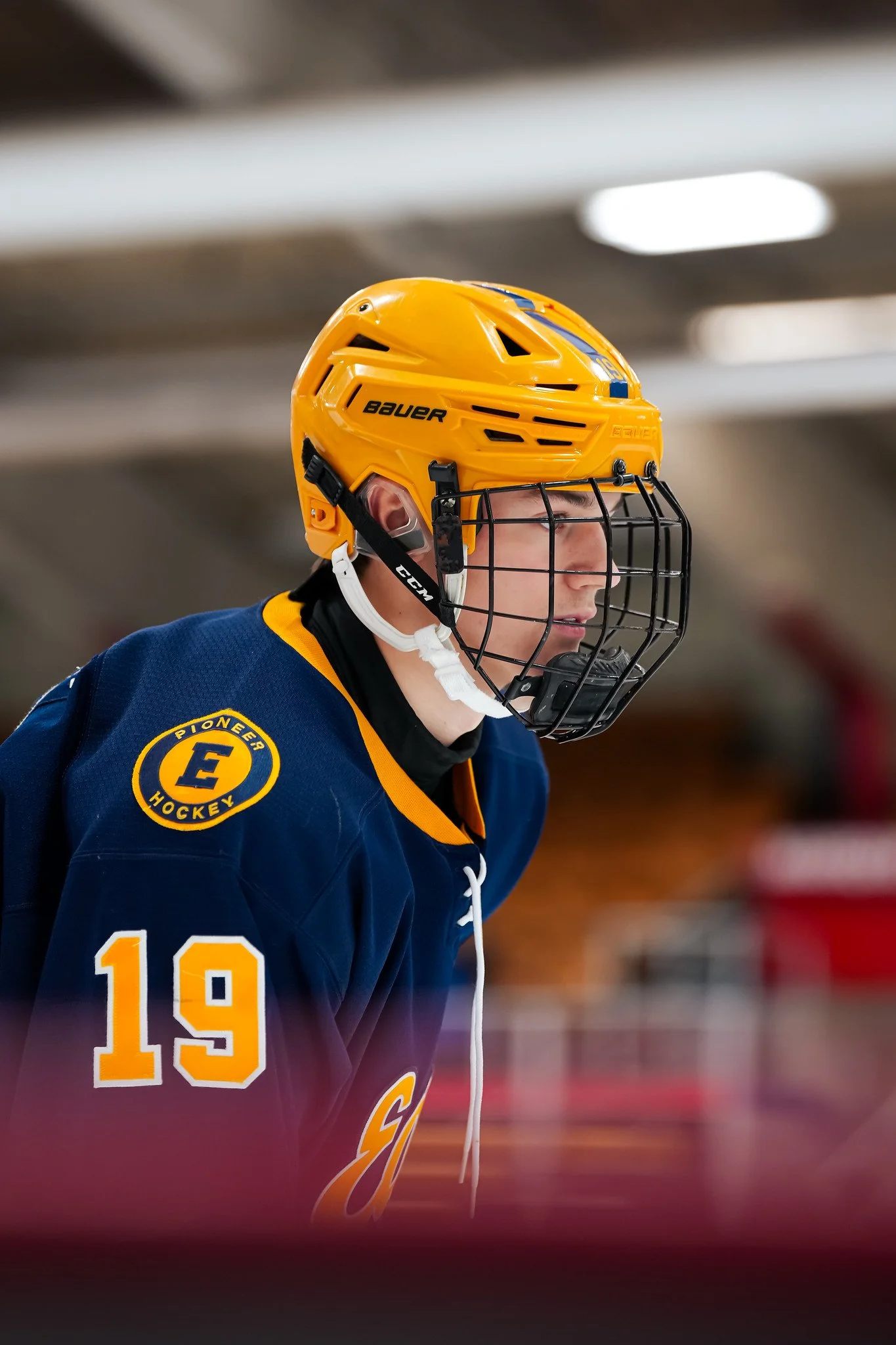 Hockey player wearing a yellow helmet, navy blue jersey, with the number 19 and an emblem on the shoulder, standing on the ice rink.