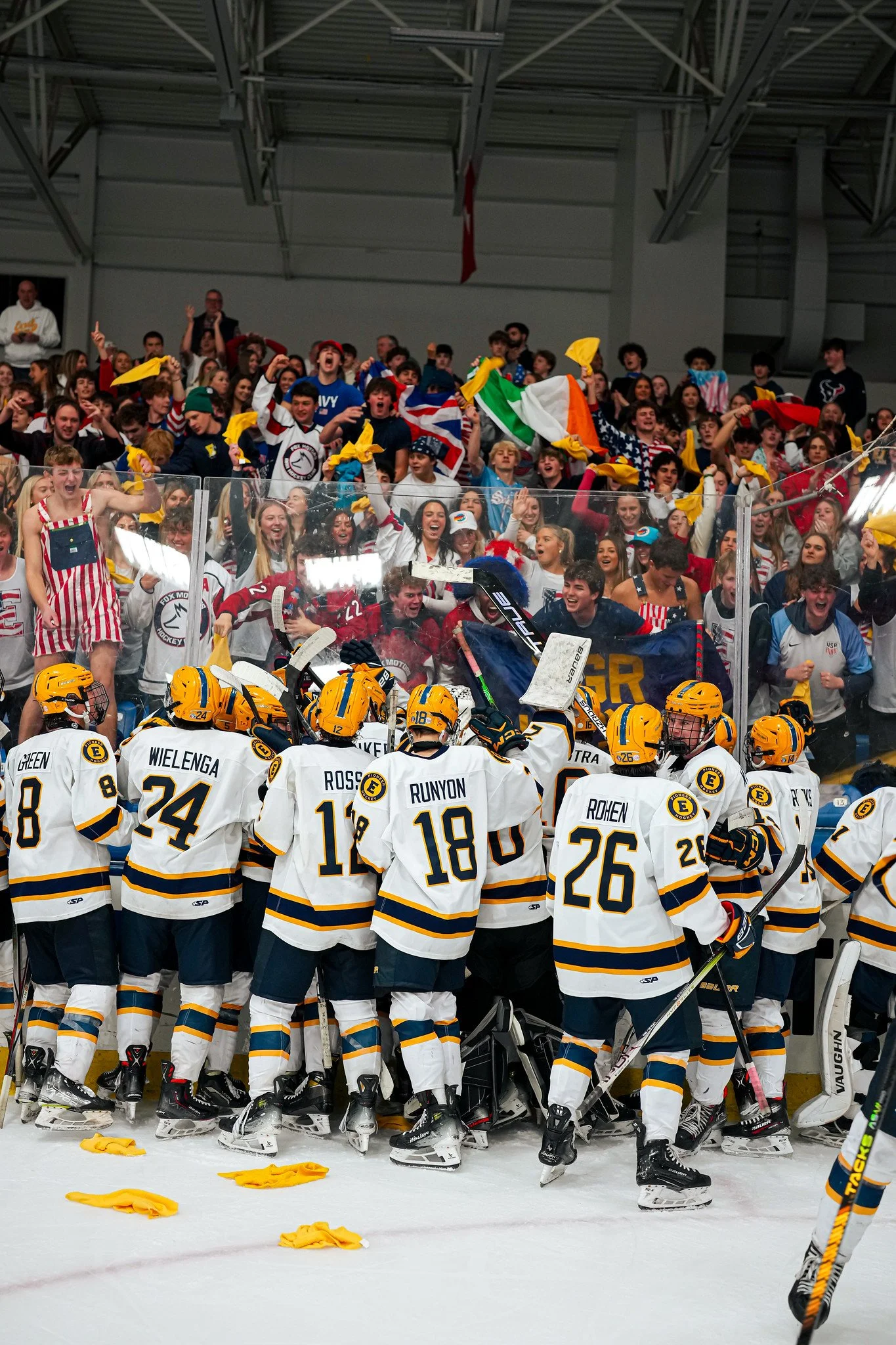 Hockey team in white and navy uniforms with yellow accents standing in a huddle on the ice rink, facing the crowd, which is cheering and waving flags.