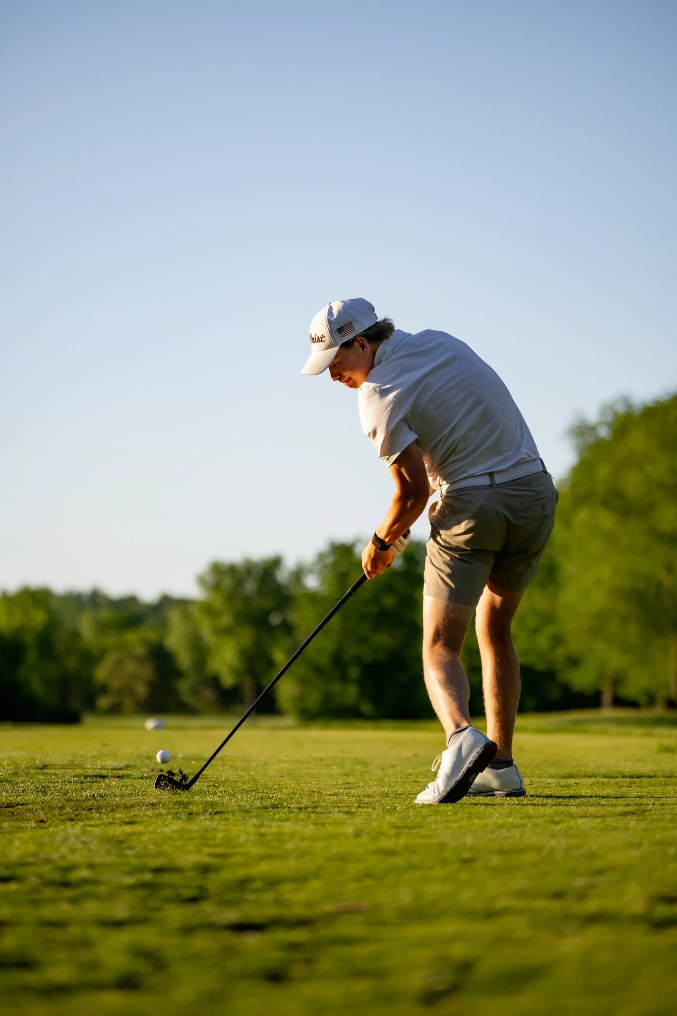 A young man wearing a white cap, gray shorts, and a light-colored shirt is playing golf on a lush green course during the late afternoon or early evening. He is about to hit a golf ball with a club, with a clear blue sky and trees in the background.