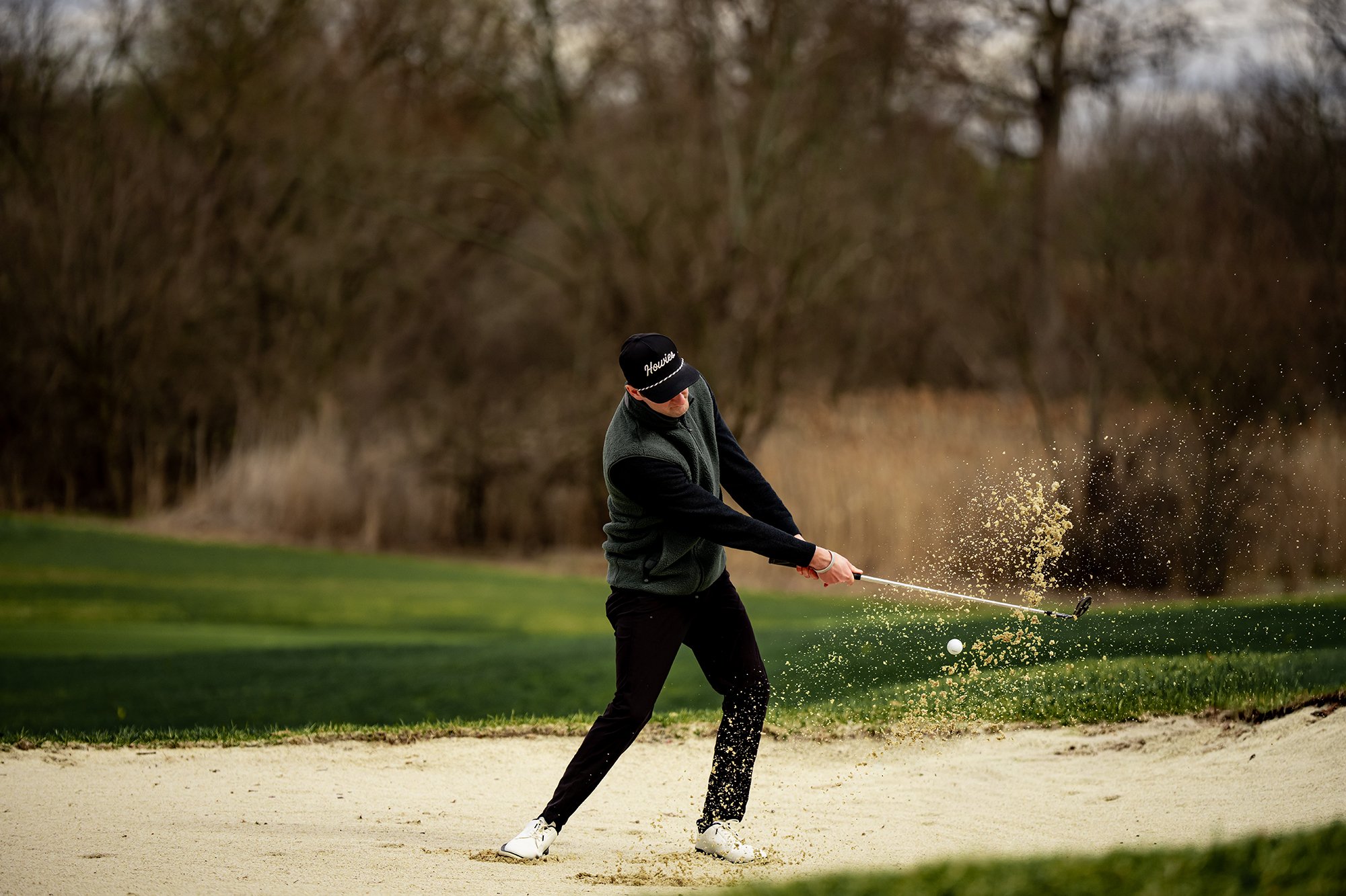 A man playing golf on a course. He is hitting a golf ball from a sand bunker, with sand flying around him. He is dressed in dark pants, a dark vest, a long sleeve shirt, and a cap.