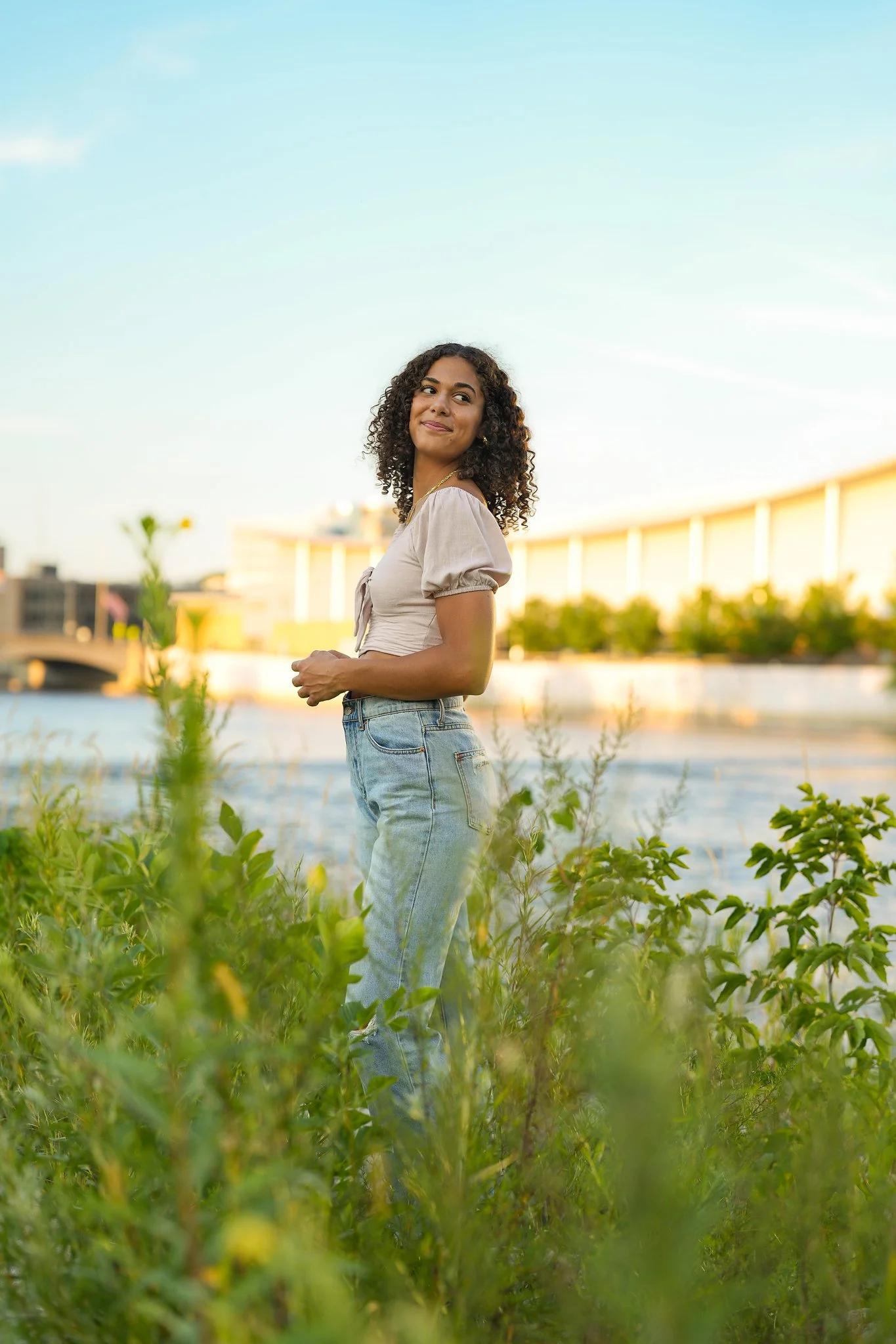 A woman standing in a green field near water during sunset, wearing a beige top and blue jeans, smiling and looking to the side.