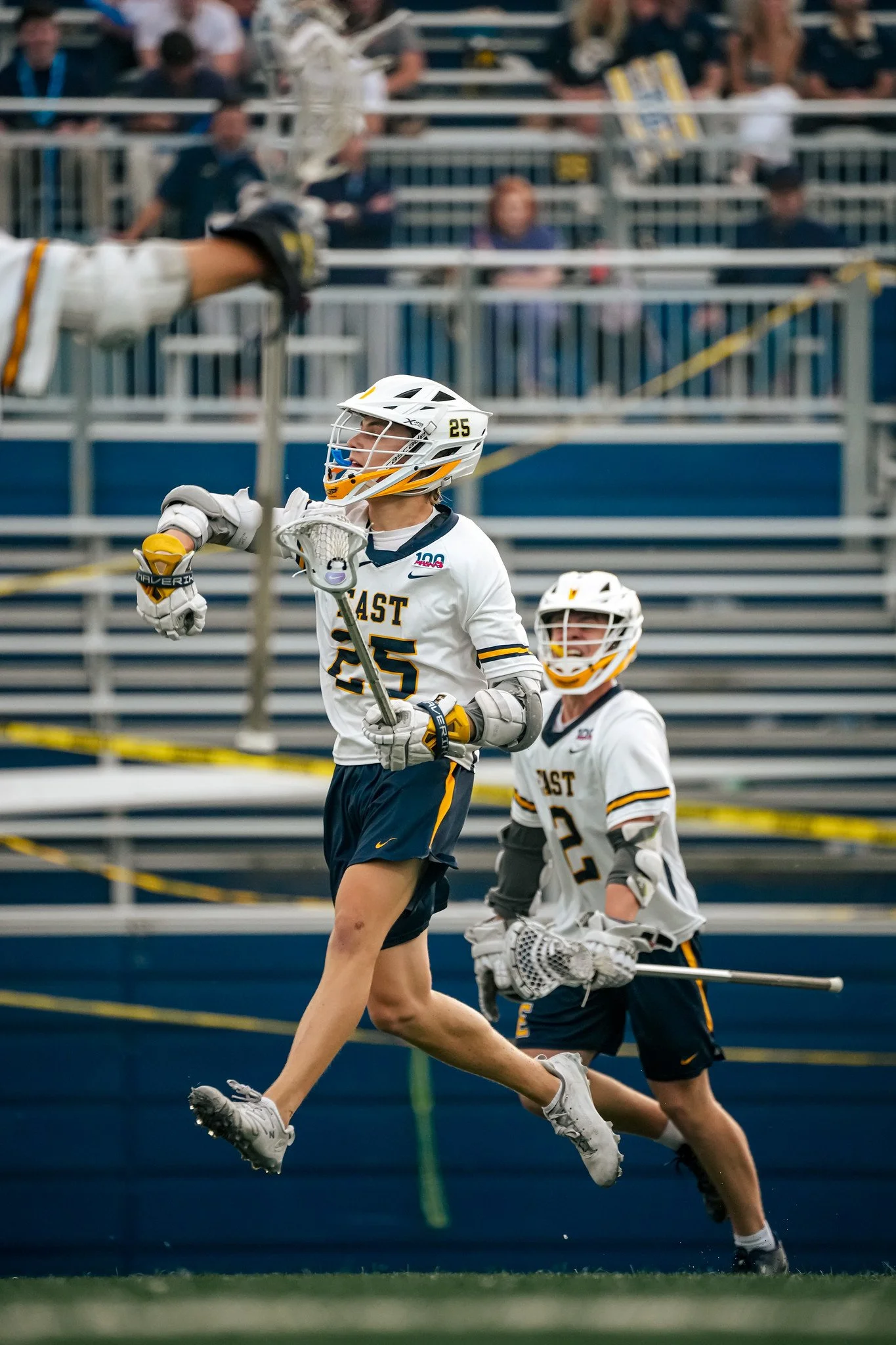 Lacrosse players in white jerseys with black and yellow accents, helmets, gloves, running across the field during a game, with spectators in the background.