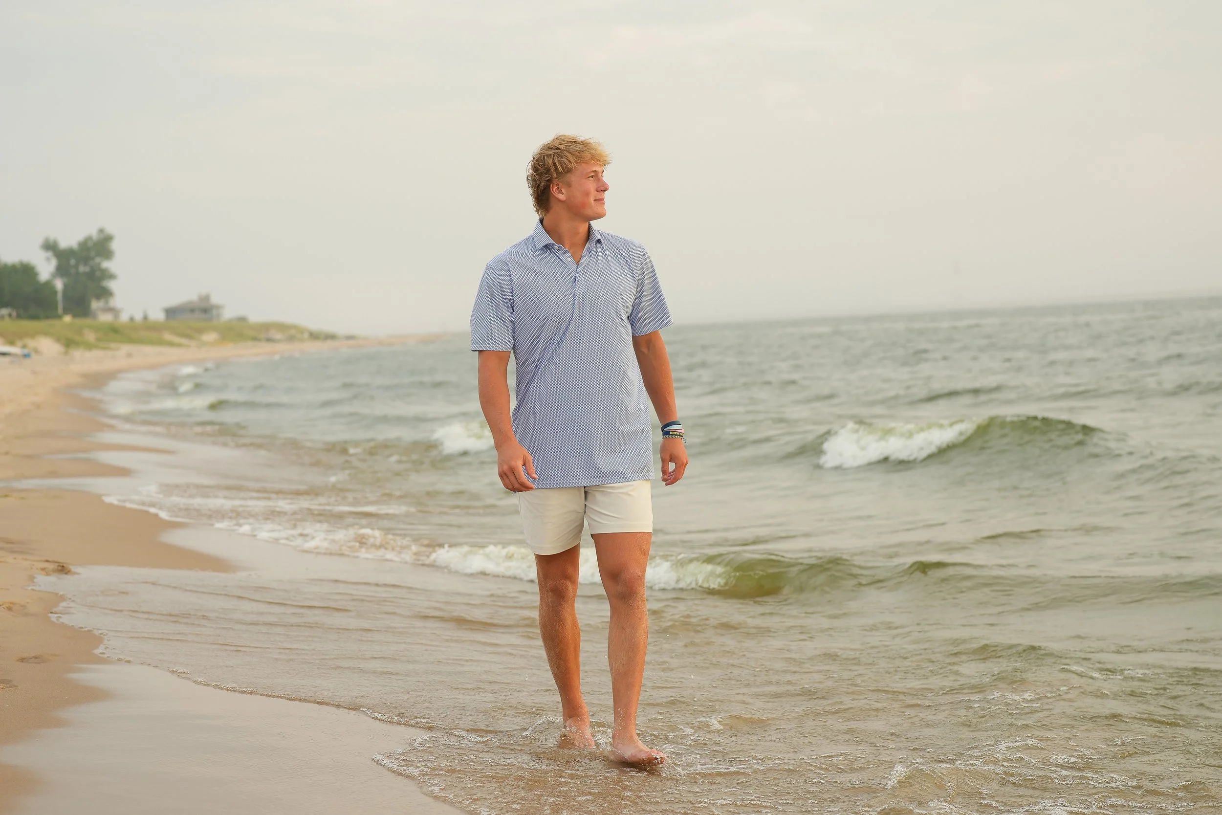 A young man walking along the shoreline at the beach with waves gently lapping at his feet, wearing a light blue short-sleeved shirt and beige shorts, with houses and trees visible in the background under a cloudy sky.