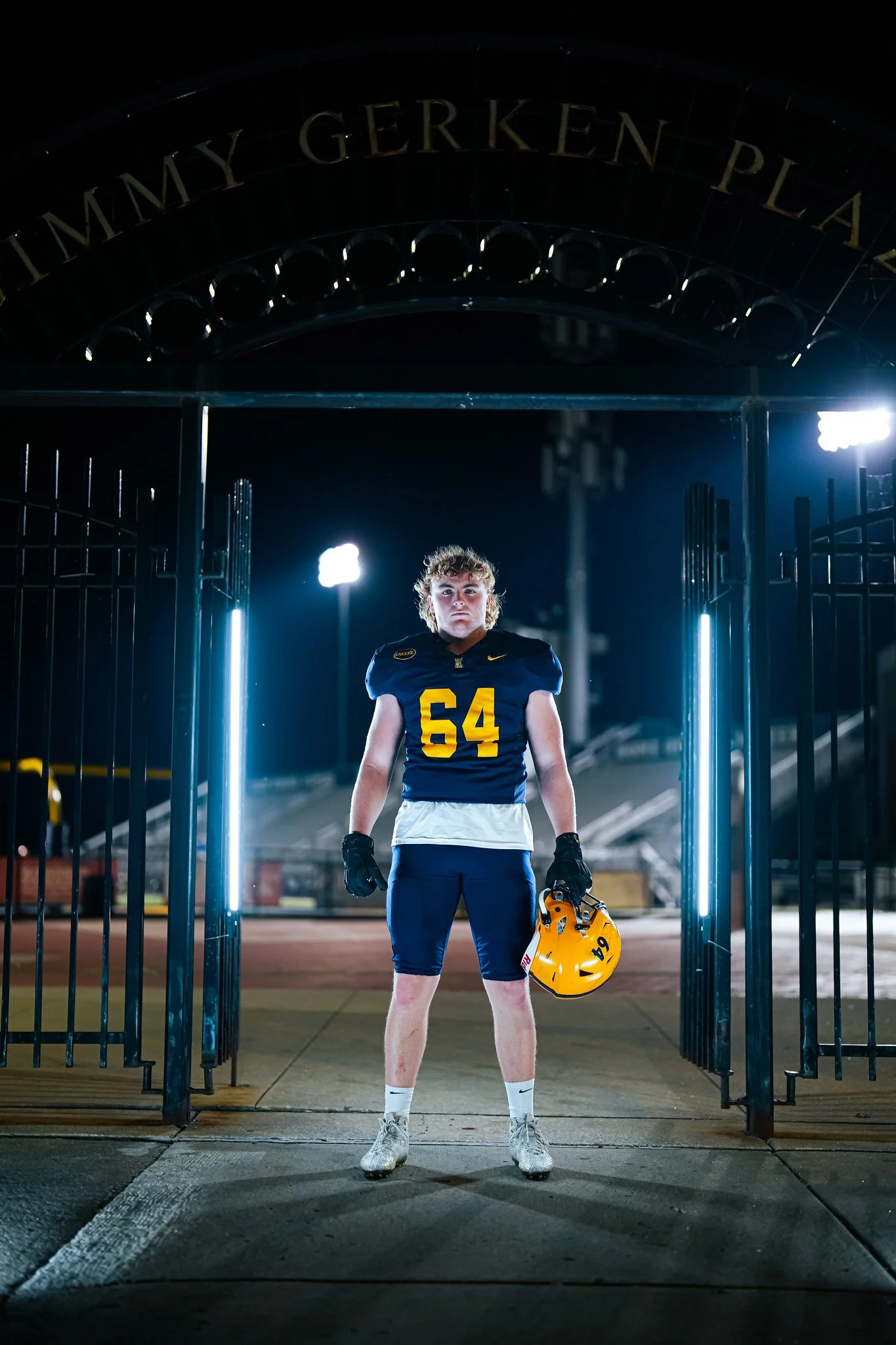 Teenage football player standing in tunnel at night, holding a yellow helmet, wearing a navy jersey with yellow number 54, white shorts, and gloves, with stadium lights in background.